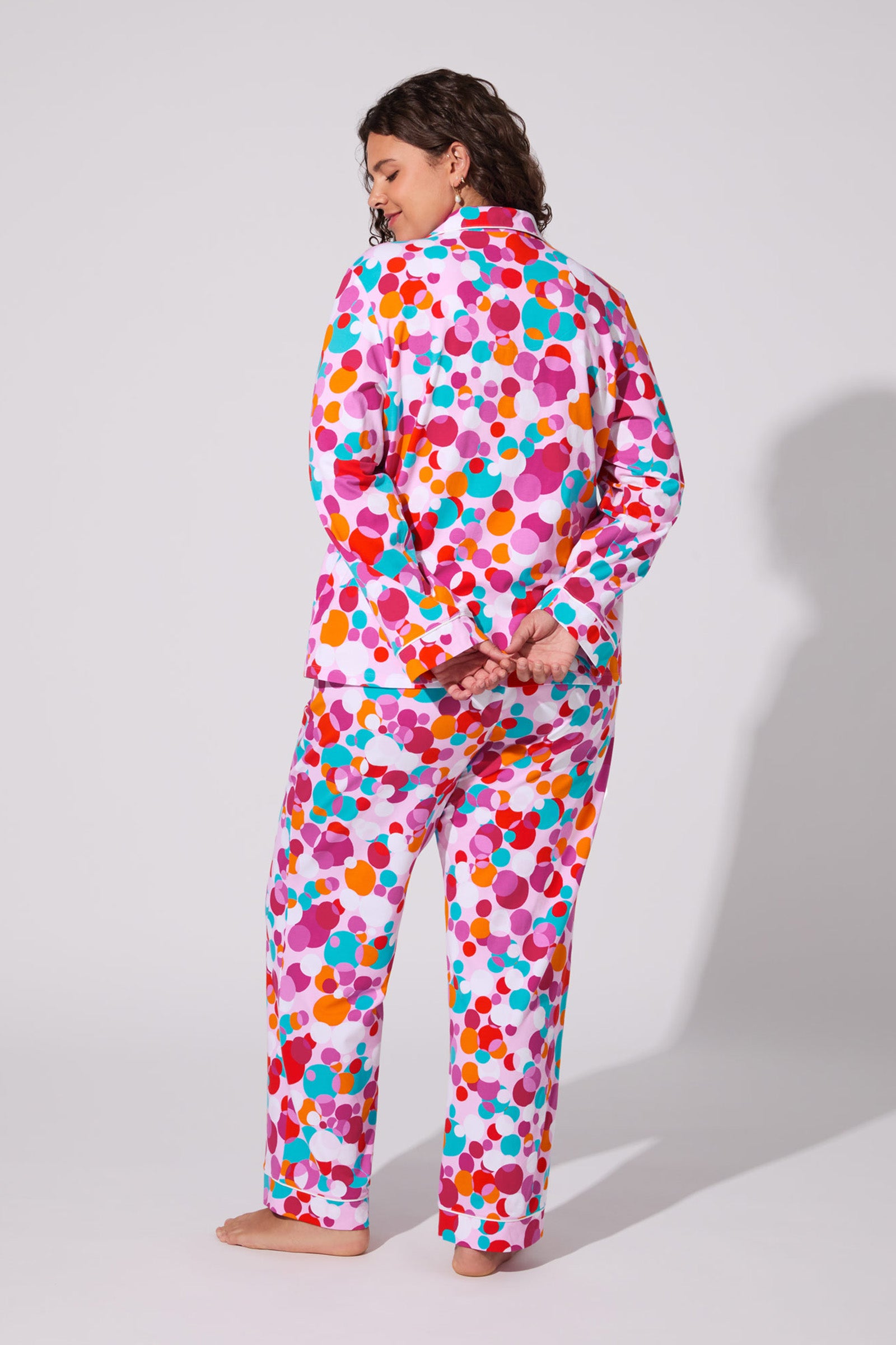 A person stands barefoot, facing away, in the Bubble Dots Classic Pj Set with colorful polka dots on a white background. Their hands are clasped behind their back, and their dark hair falls in loose waves.