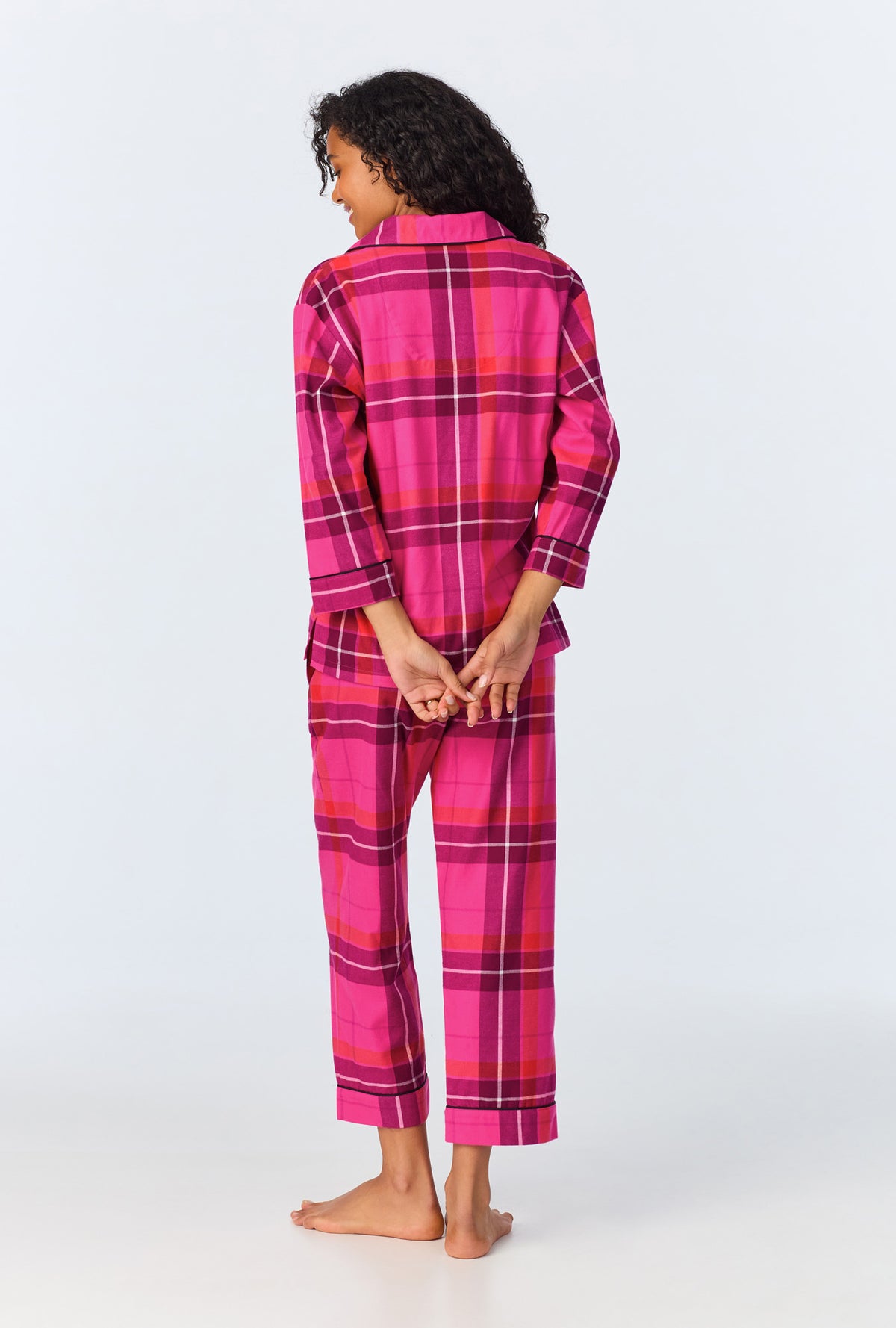 A person stands barefoot facing away, wearing the Aurora Plaid Womens 3/4-Sleeve Cropped Pant Flannel PJ Set in pink and magenta. Their hands are clasped behind their back, and they have curly hair.