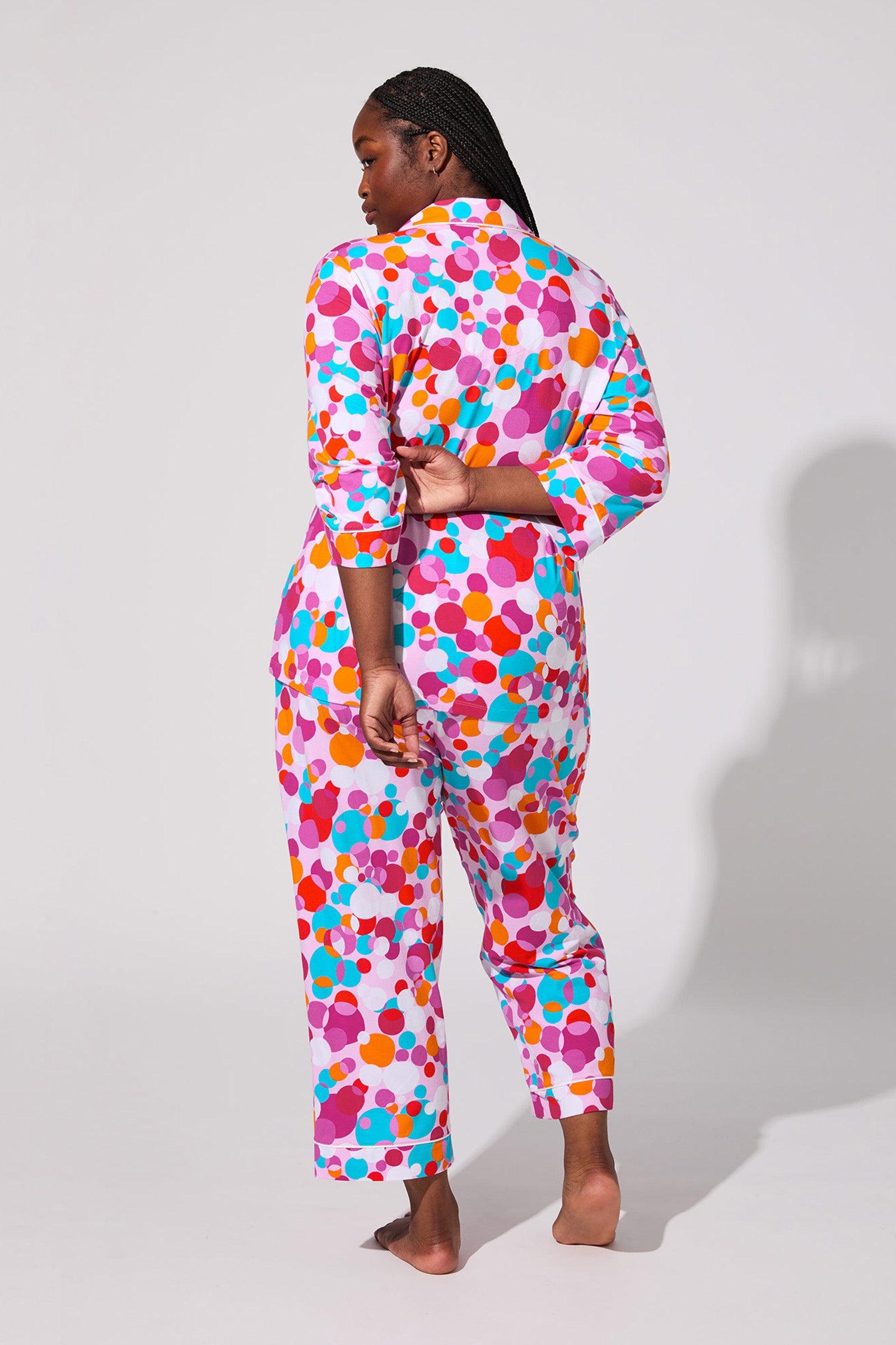 A person with braided hair stands barefoot on a light background, facing away, wearing the Bubble Dots Crop Pj Set featuring colorful polka dots in pink, purple, orange, red, and blue.