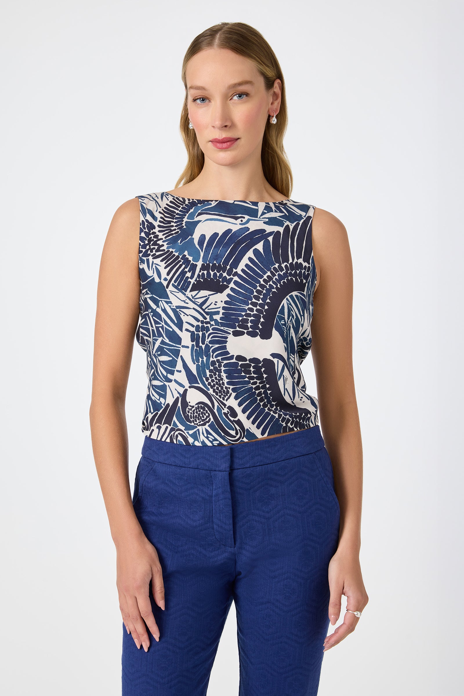 A woman wears the Hakuto Tie-Back Top in Tsuru, featuring a blue and white abstract bird pattern, paired with blue textured pants. She stands against a plain white background, facing forward with a slight smile.