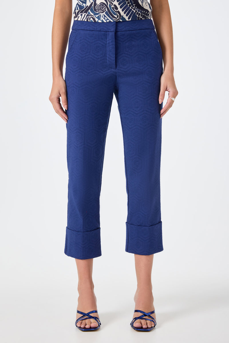 A woman wears Banshee Cropped Cuff Hem Pants in Noh Nightfall, styled with a patterned navy and white top and blue strappy sandals, standing against a plain white background.