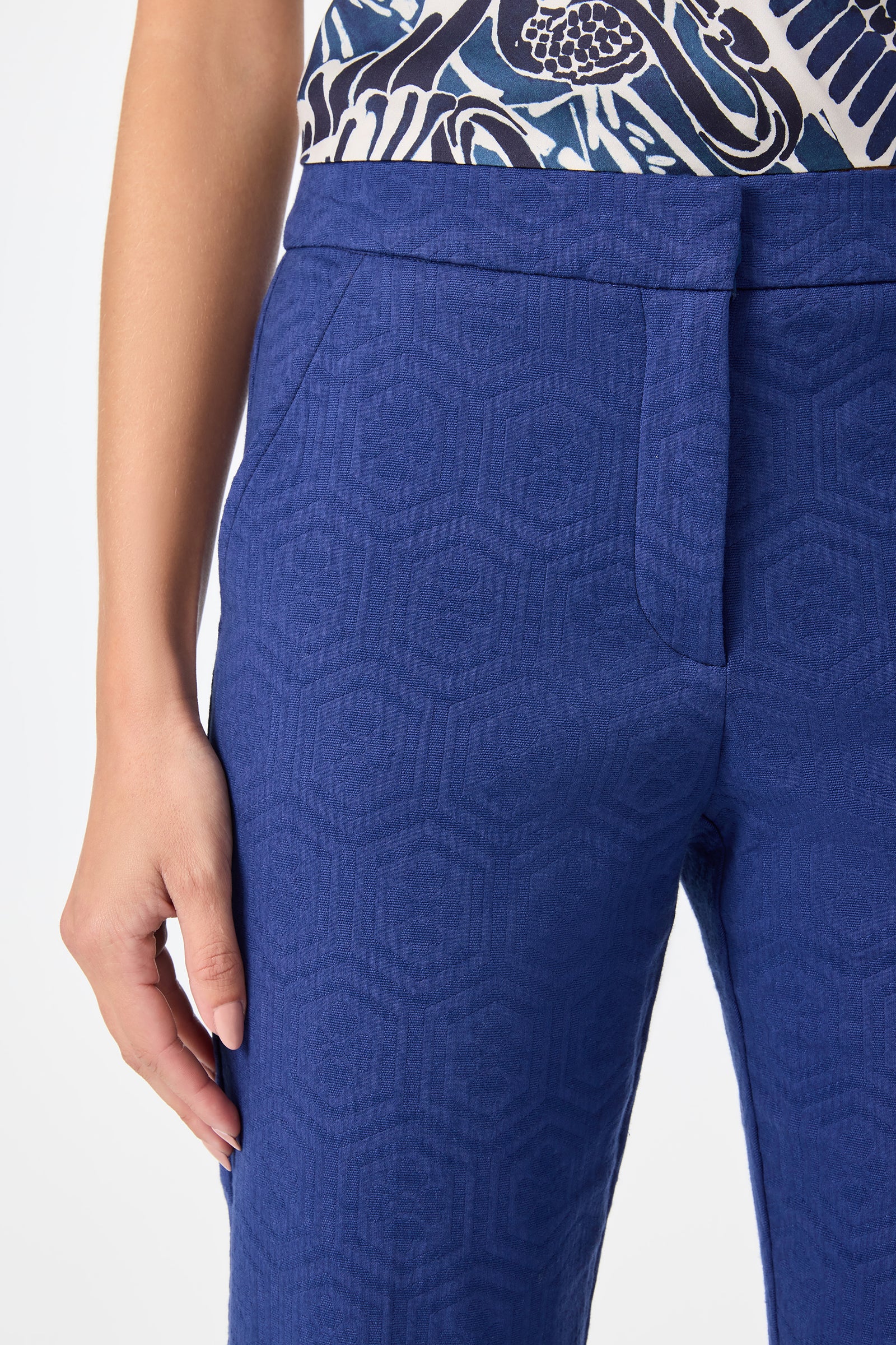 A person wearing Banshee Cropped Cuff Hem Pants in textured blue with a subtle geometric pattern and a dark blue and white patterned top, their left hand resting by their side.