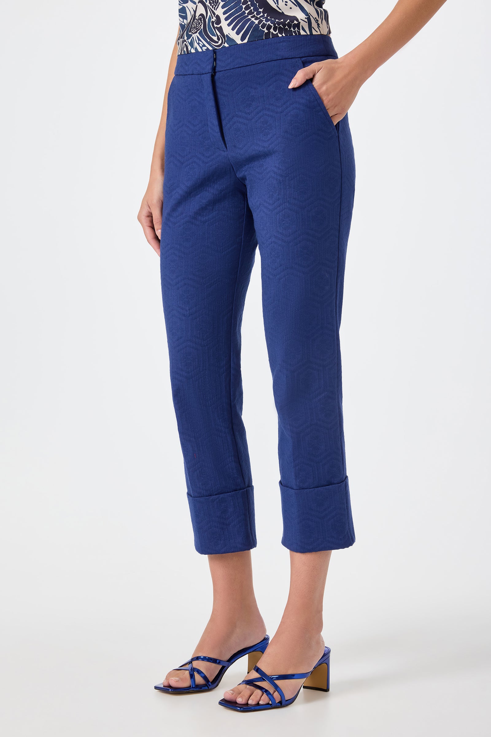 A person stands against a plain background, hands in pockets, wearing Banshee Cropped Cuff Hem Pants in royal blue with a geometric texture, paired with a patterned top and blue strappy block-heeled sandals.