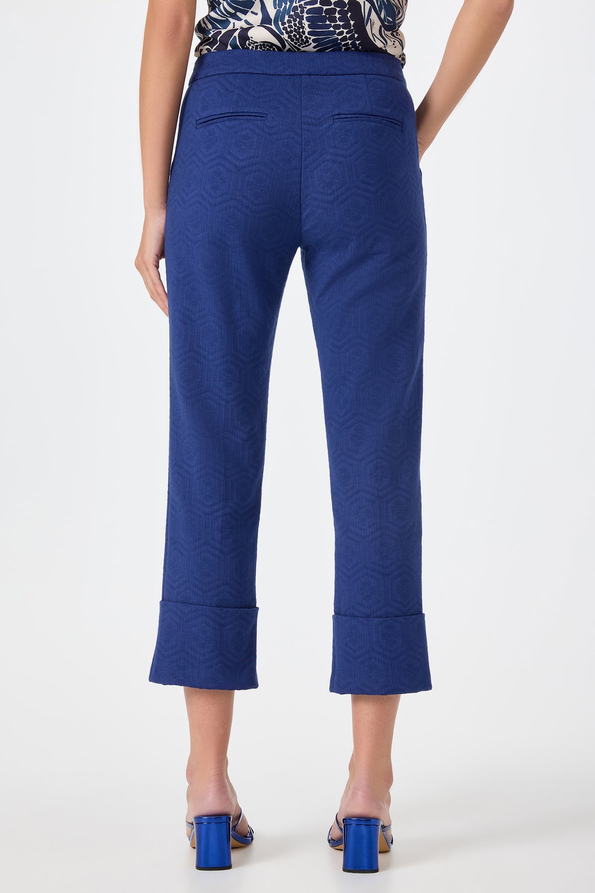 A person faces away wearing Banshee Cropped Cuff Hem Pants with a geometric pattern, cuffed hems, and blue high-heeled sandals against a plain white background.