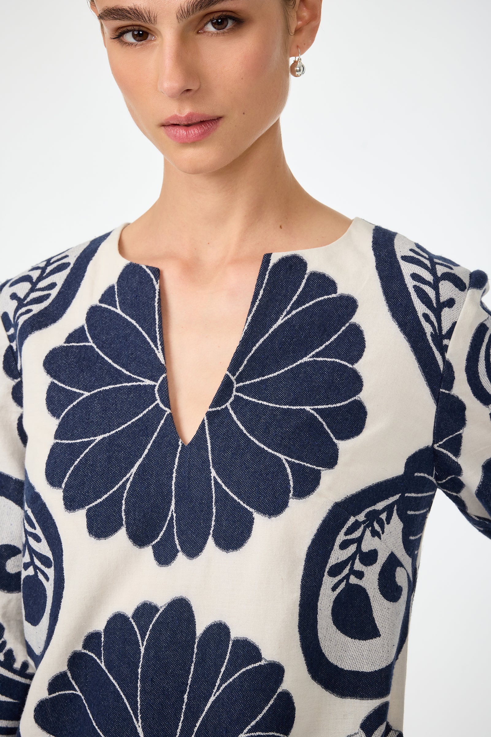 A woman wearing the Amy 3/4 Sleeve Dress - Fuyuko Fil Coupe, featuring a cream base with bold navy blue floral patterns and a deep V neckline, looks slightly past the camera against a plain light background.