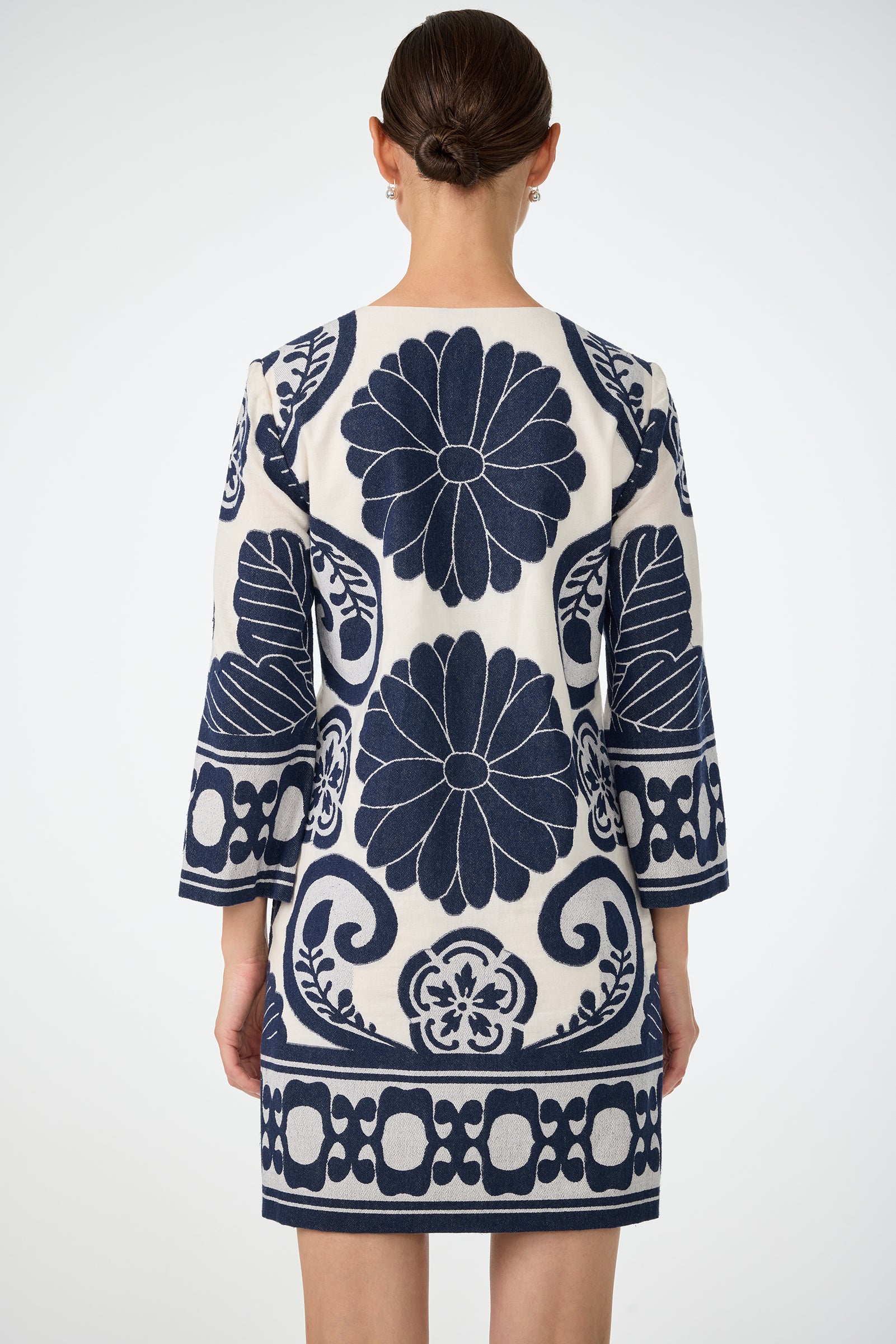 A woman with her hair in a low bun is seen from the back, wearing the Amy 3/4 Sleeve Dress - Fuyuko Fil Coupe, a white dress featuring bold navy blue floral and geometric patterns against a plain light background.