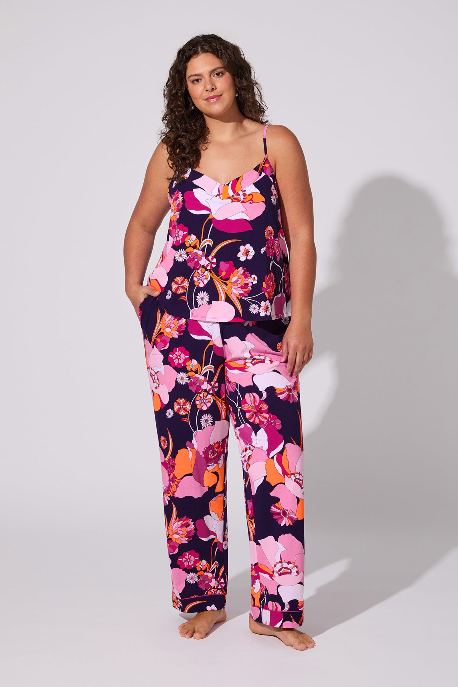 A woman with long curly hair models the Floral Medley Cami PJ Set, showcasing its vibrant pink, purple, and orange floral pattern as she stands barefoot against a plain light background.