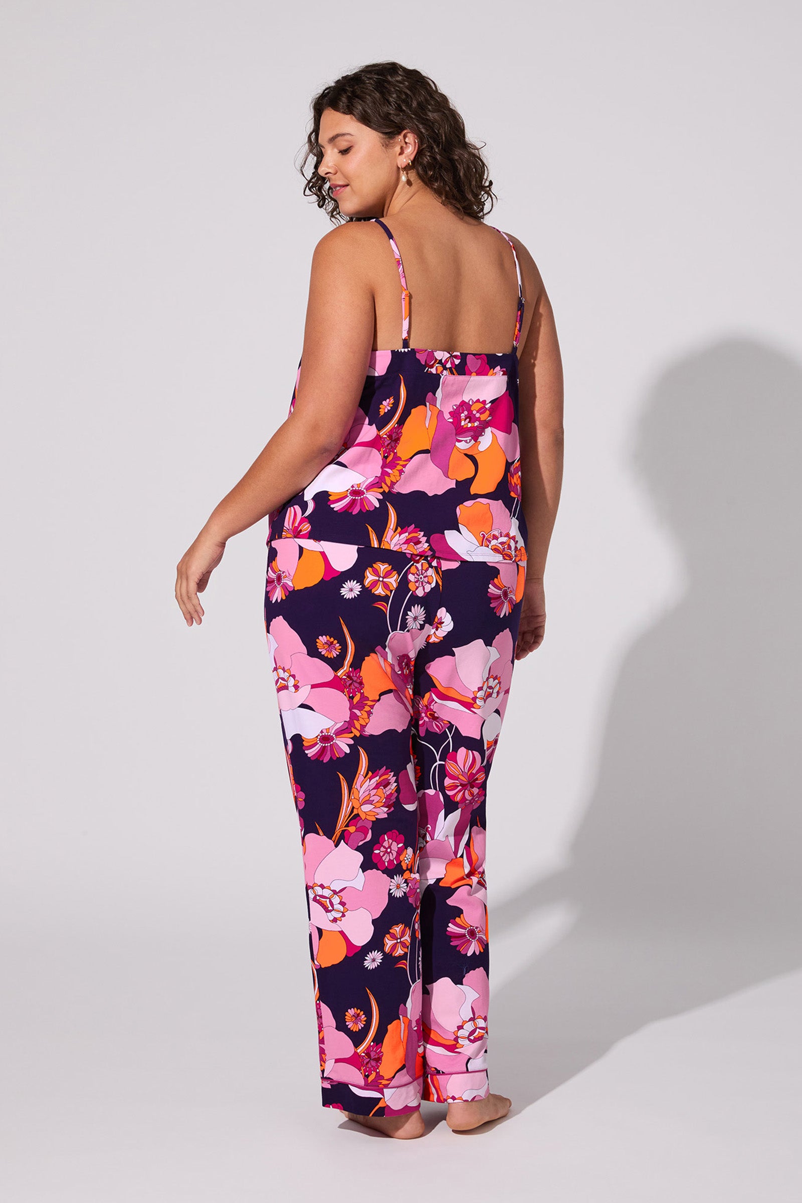 A woman with curly hair stands barefoot, facing away, wearing the Floral Medley Cami Pj Set—a colorful floral print pajama outfit with a cami top and pants in shades of pink, purple, and orange against a plain light background.