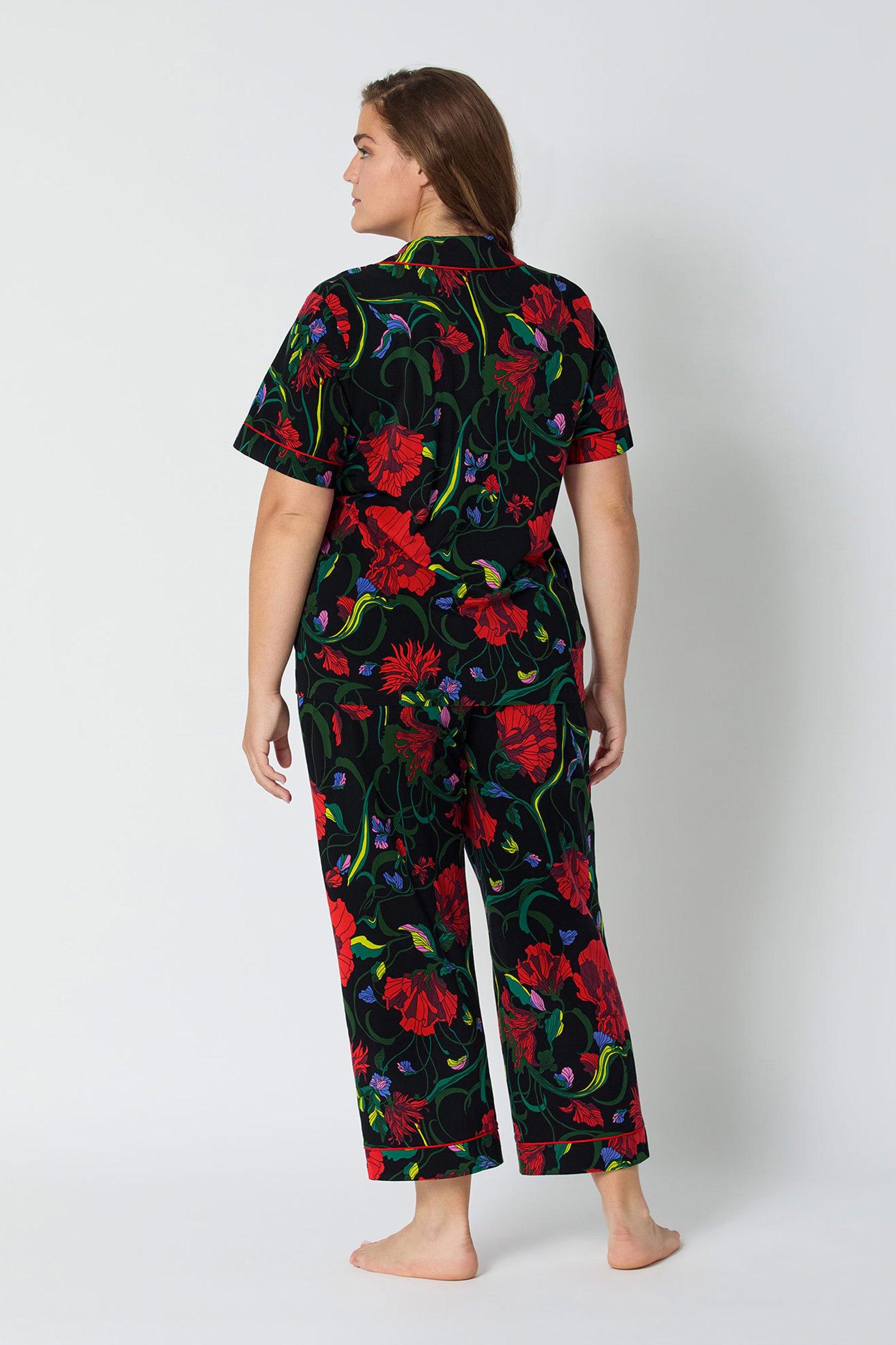 A woman faces away barefoot, wearing the Haute Floral S/S Cropped PJ Set—a matching short-sleeve top and long pants featuring a bold red and green floral pattern on a black background.