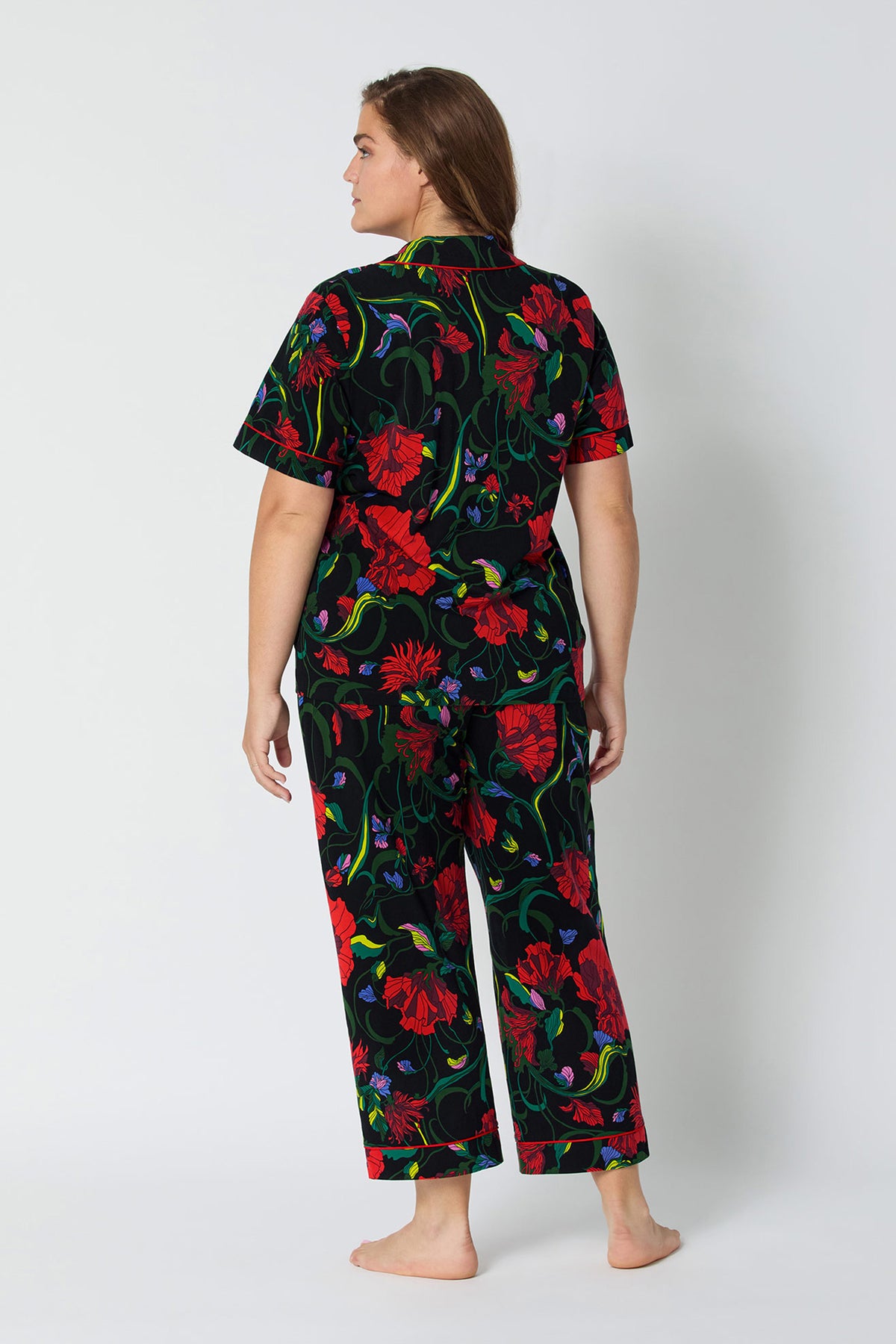 A woman faces away barefoot, wearing the Haute Floral S/S Cropped PJ Set—a matching short-sleeve top and long pants featuring a bold red and green floral pattern on a black background.