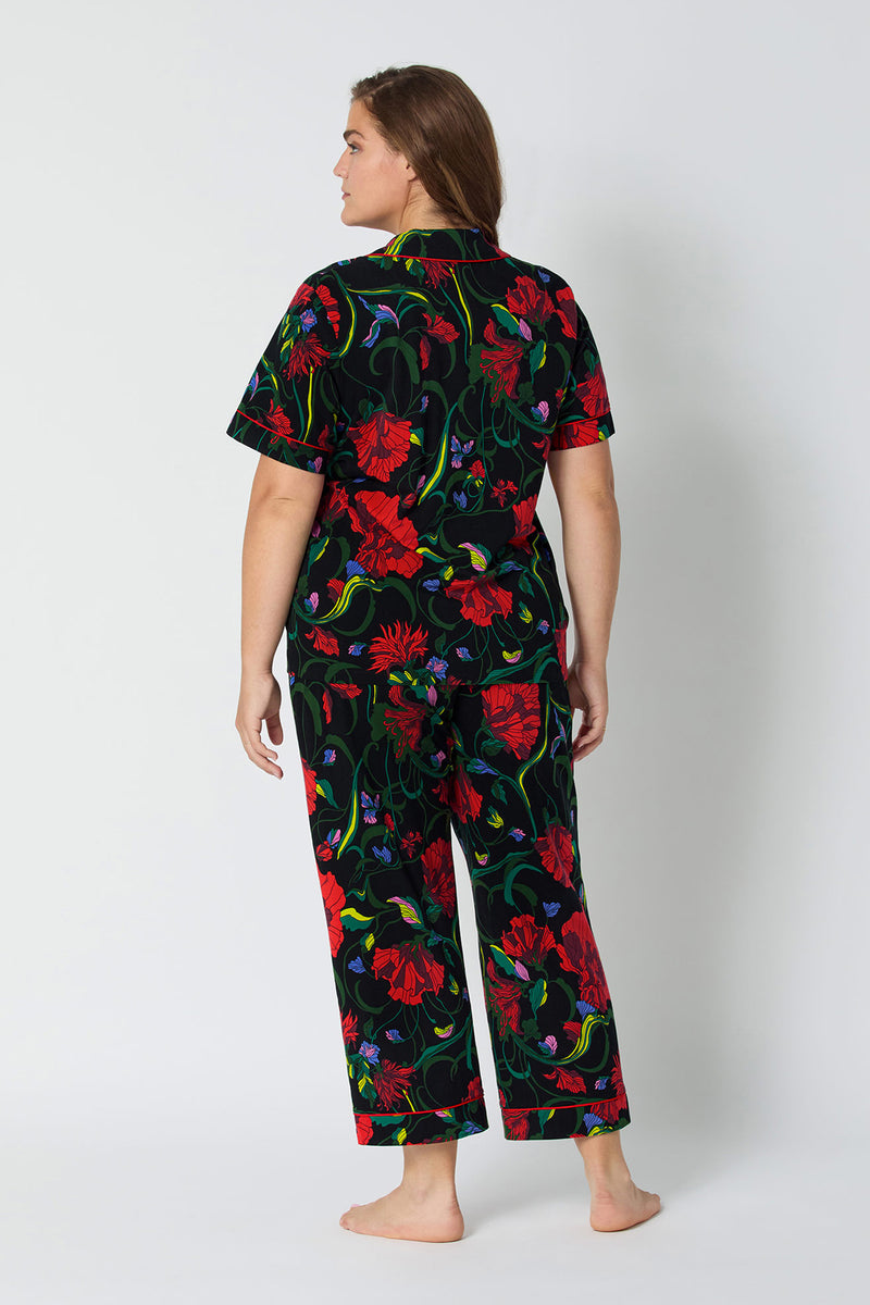 A woman faces away barefoot, wearing the Haute Floral S/S Cropped PJ Set—a matching short-sleeve top and long pants featuring a bold red and green floral pattern on a black background.