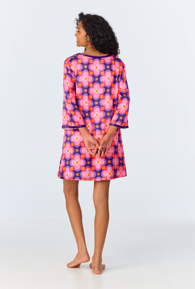 A woman with curly dark hair stands barefoot facing away, wearing the Space Cat Womens 3/4-Length Sleeve Silk Caftan—a knee-length, washable silk dress featuring bold pink, orange, and purple floral patterns. Her arms rest behind her back.
