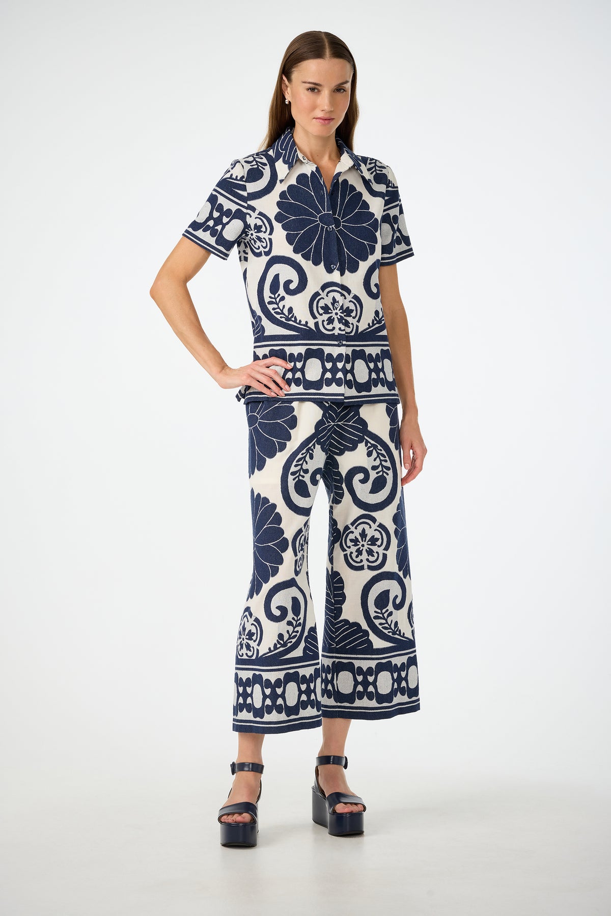 A woman wears the Estella 2 Cropped Pants - Fuyuko Fil Coupe, a matching short-sleeve top and wide-leg set featuring bold navy blue floral and swirl patterns on white, styled with black platform sandals.