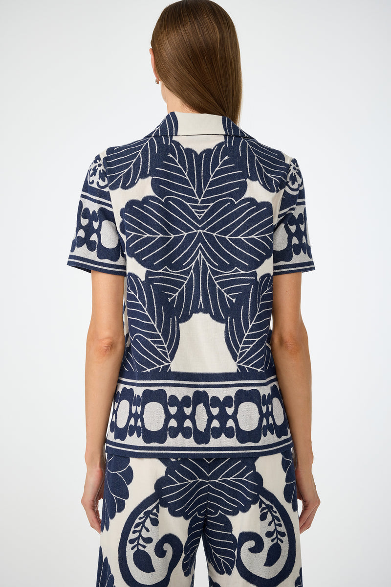Someone with long brown hair, seen from behind, wears the Abba Top - Fuyuko Fil Coupe, a short-sleeve shirt with bold navy blue and white floral and geometric patterns, paired with matching pants.