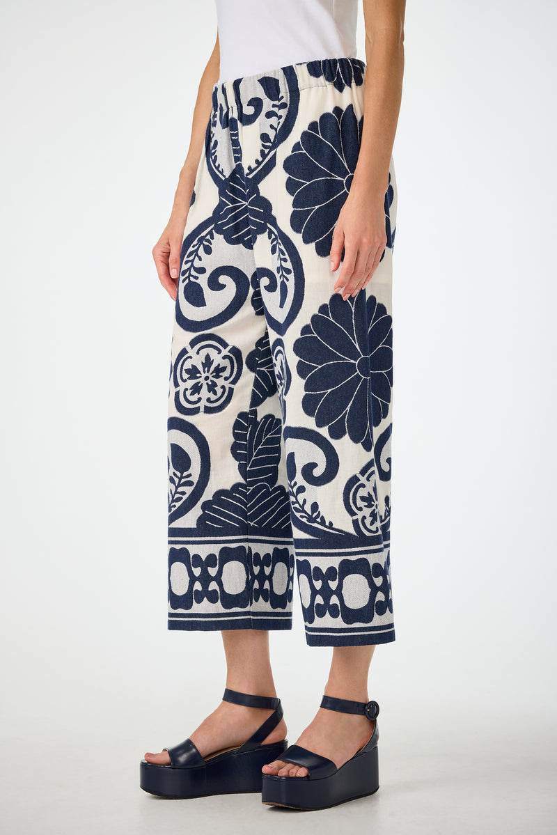A person wears the Estella 2 Cropped Pants - Fuyuko Fil Coupe, featuring wide-leg, cropped white pants with navy floral and geometric patterns, paired with a white top and black platform sandals against a plain white background.