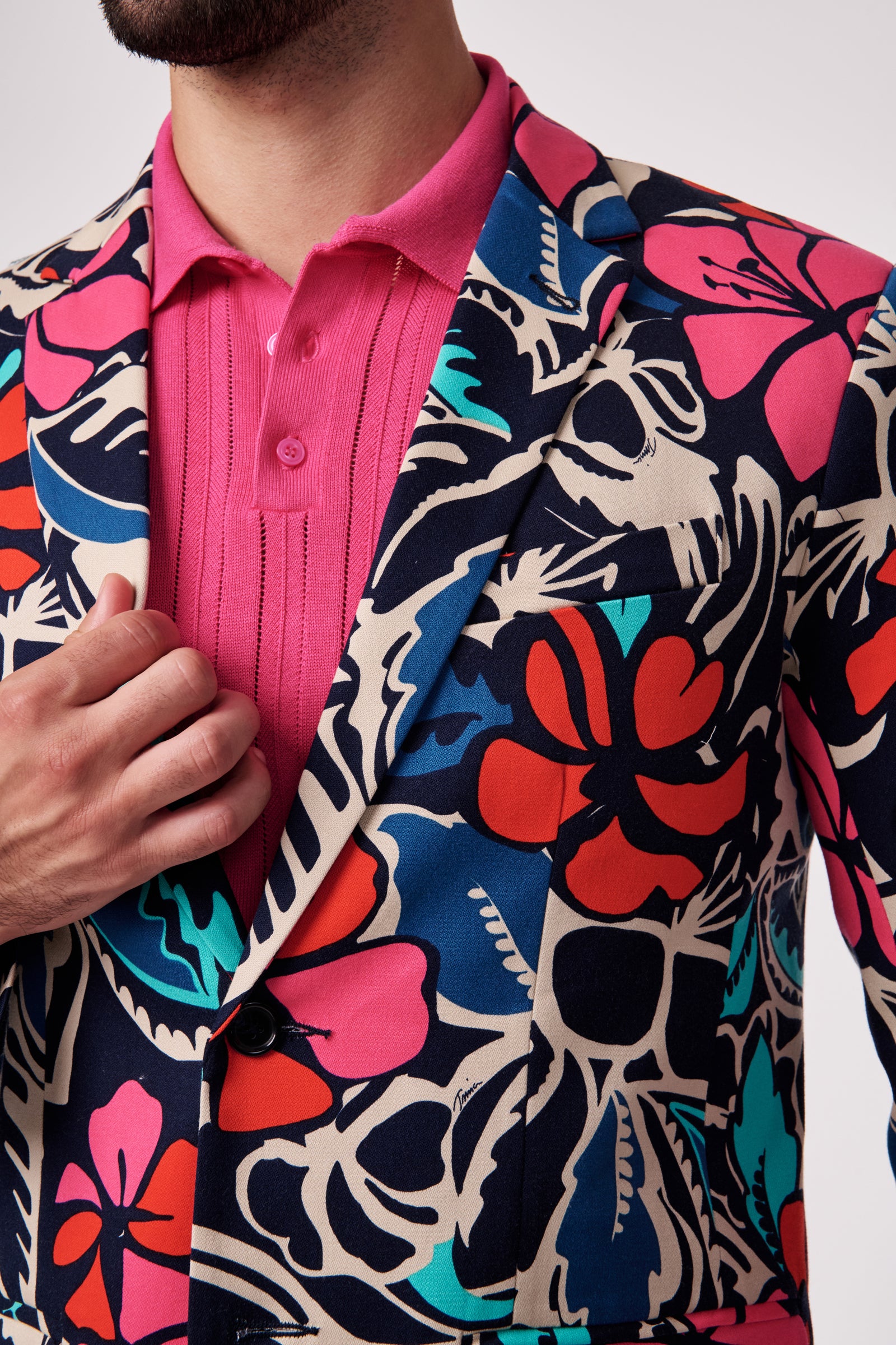 Wearing the Thurston Blazer, featuring bold floral patterns in red, pink, blue, teal, and cream over a bright pink collared shirt, a person adjusts the blazer with their left hand.