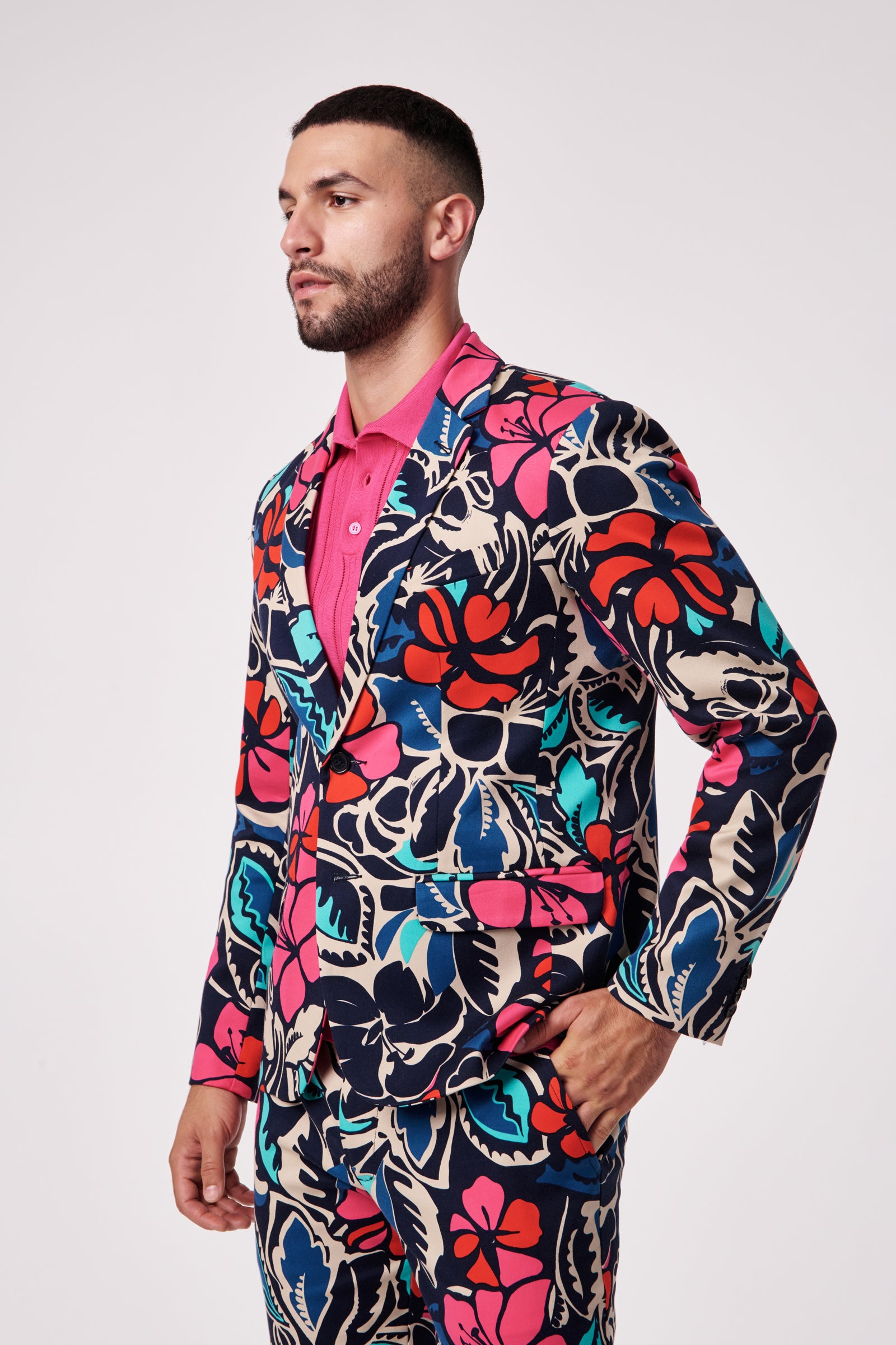 A man wears the Thurston Blazer, featuring a bold tropical floral pattern in red, blue, teal, and cream, paired with a pink shirt against a plain light background.