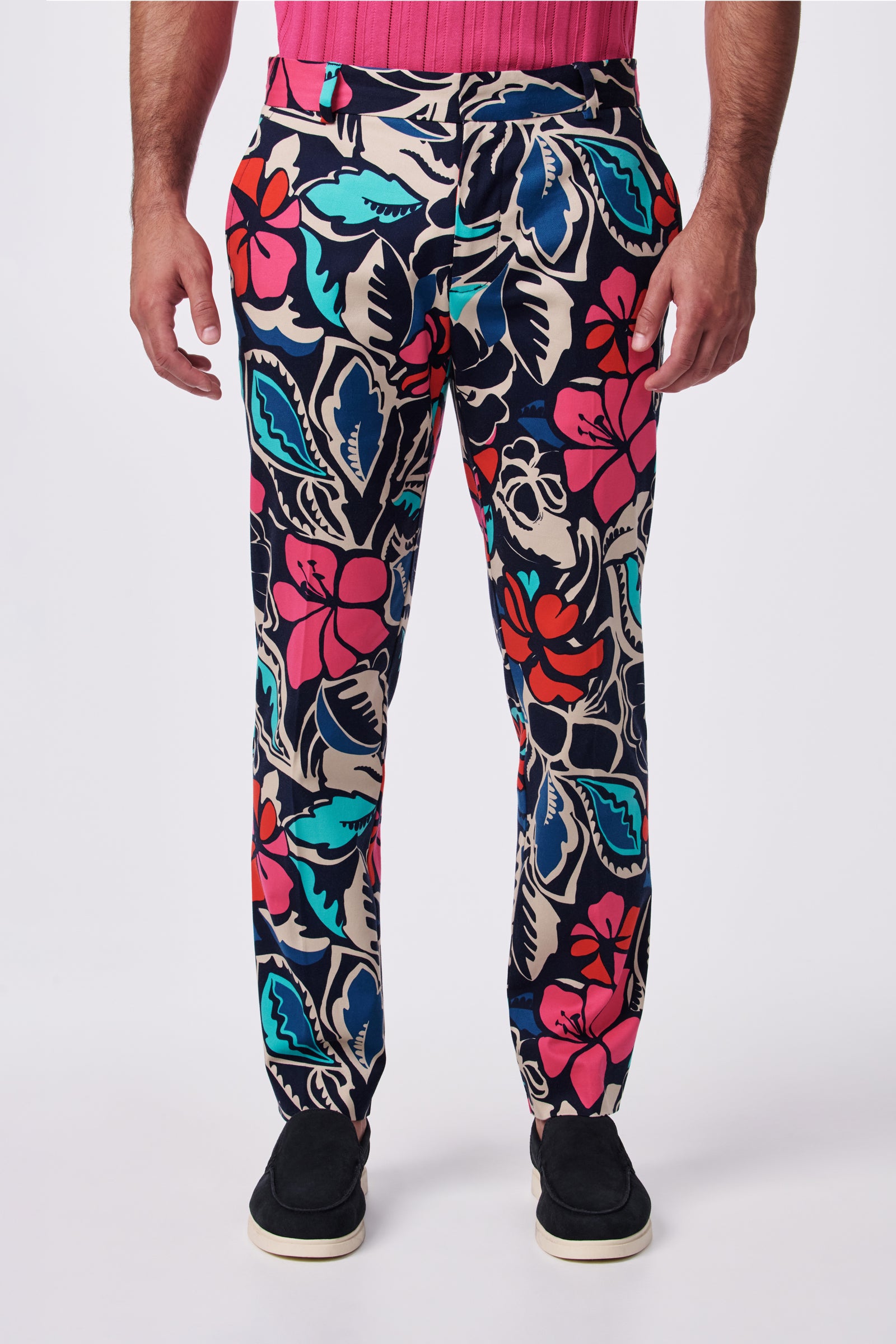A person wearing Clyde Slim Trouser with a bold floral pattern in red, blue, pink, and beige, paired with a pink shirt and black slip-on shoes, photographed against a white background.