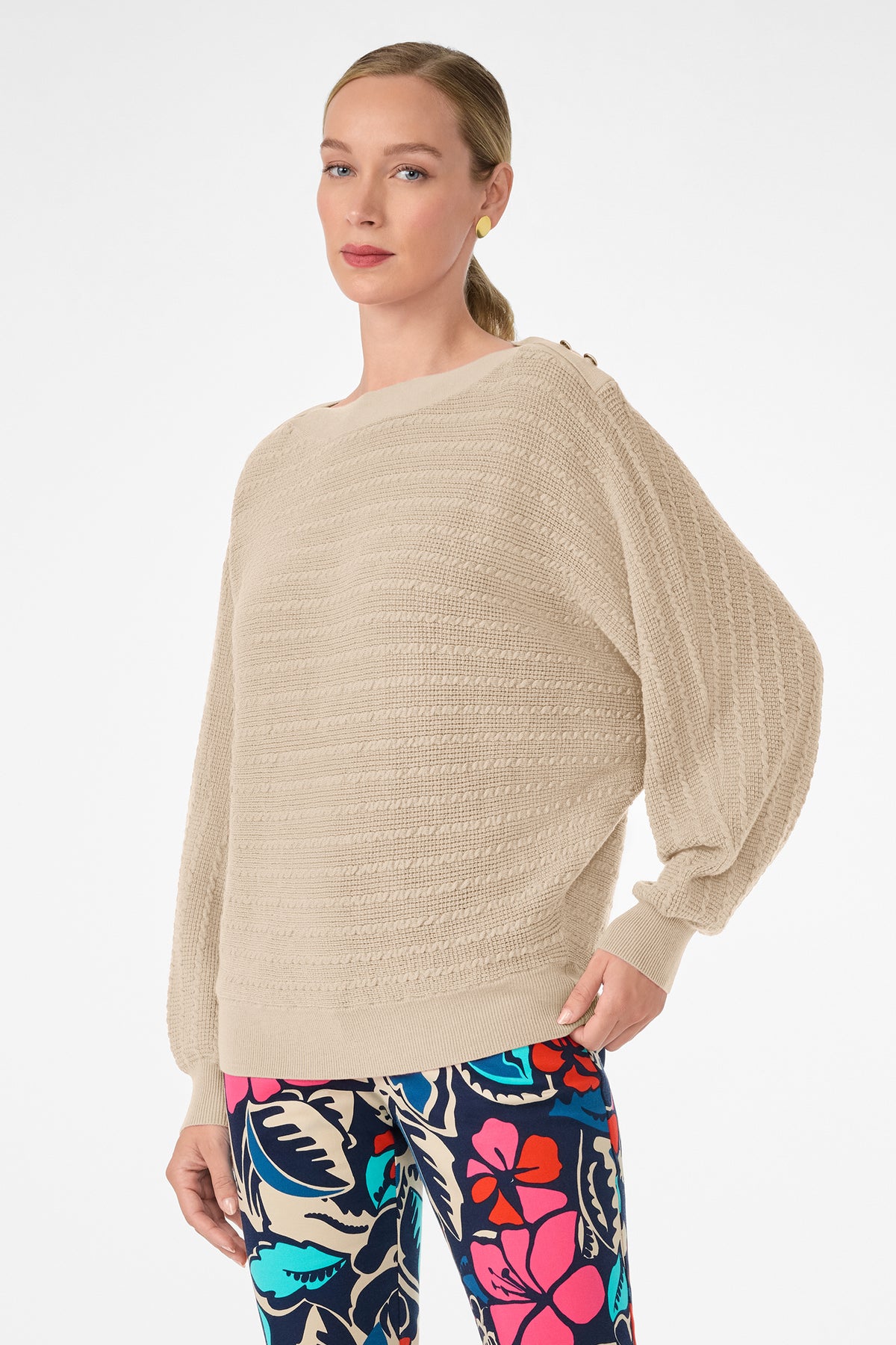 A light-skinned, blonde-haired woman with a low ponytail wears the Ima Sweater, a loose beige textured knit, paired with colorful floral leggings. She stands against a plain white background, looking slightly to the side.