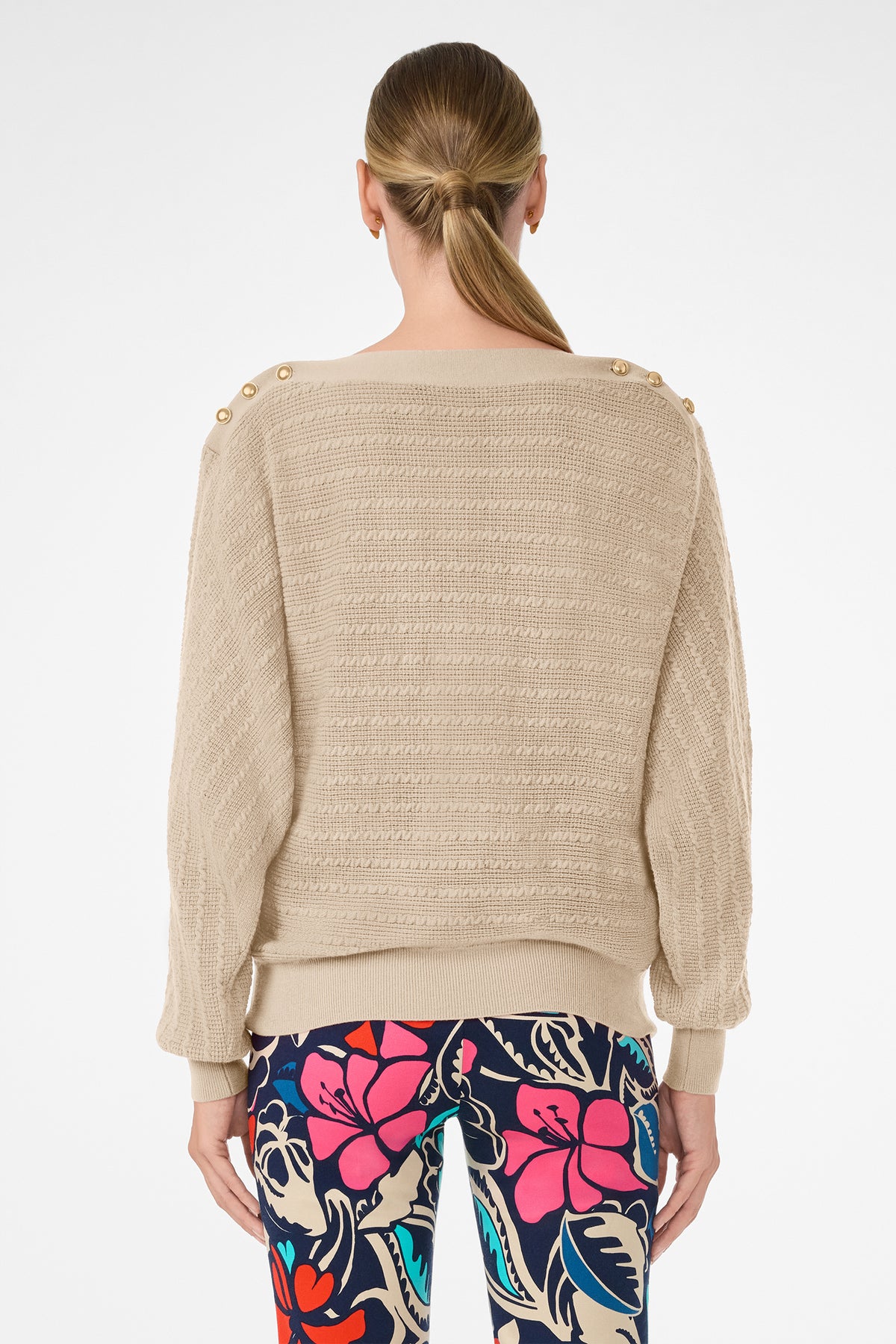 A woman with straight, light brown hair in a low ponytail is shown from the back wearing the Ima Sweater—a beige textured knit with gold shoulder buttons—paired with colorful floral pants against a plain white background.