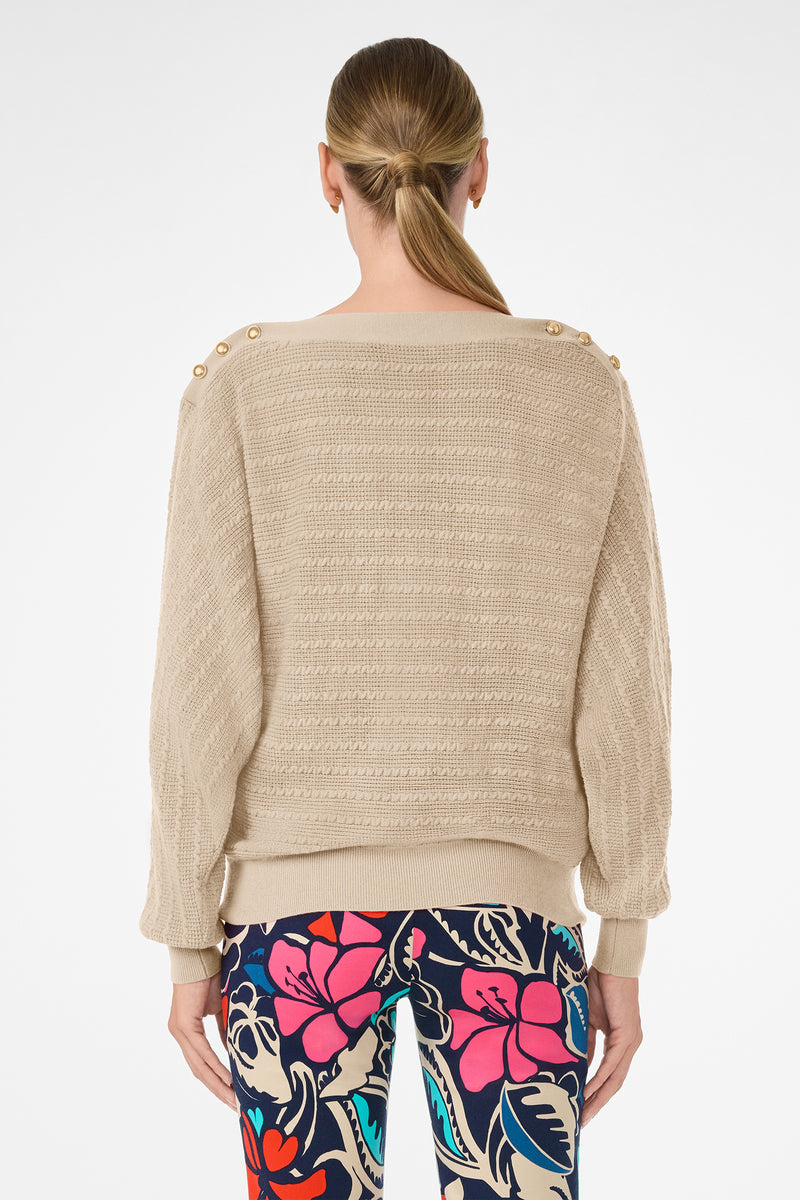 A woman with straight, light brown hair in a low ponytail is shown from the back wearing the Ima Sweater—a beige textured knit with gold shoulder buttons—paired with colorful floral pants against a plain white background.