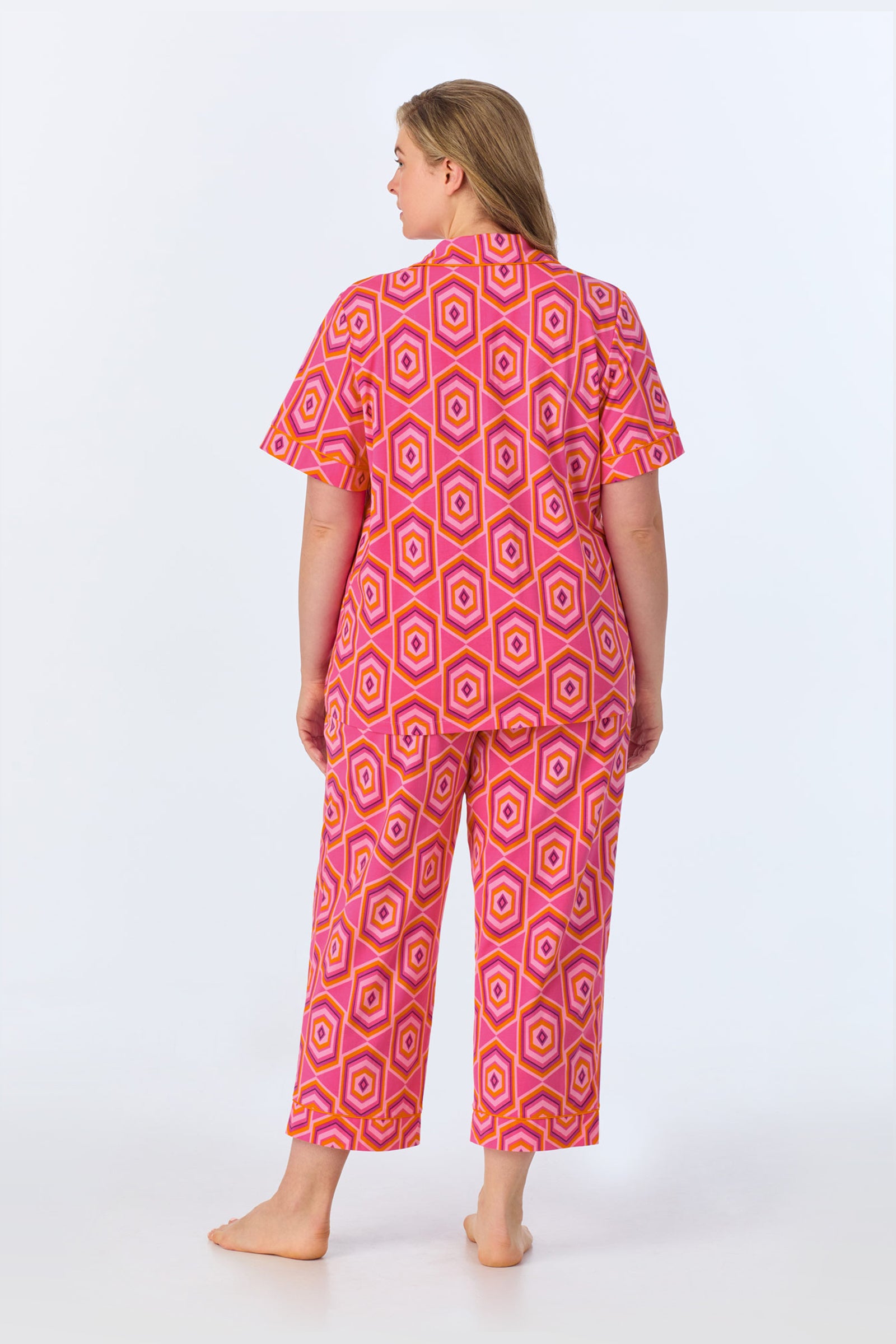 A woman stands barefoot facing away, wearing the TT SS Crop PJ Set - MLT Hexagon Jersey with bright pink, orange, and yellow geometric hexagon patterns against a plain white background.