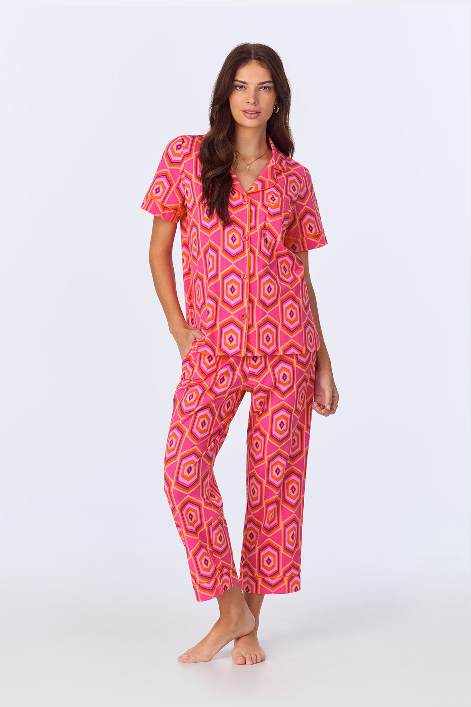 A woman stands barefoot against a white background wearing the TT SS Crop PJ Set - MLT Hexagon Jersey in pink, featuring a bold geometric pattern, short sleeves, a collar, and one hand in her pocket.