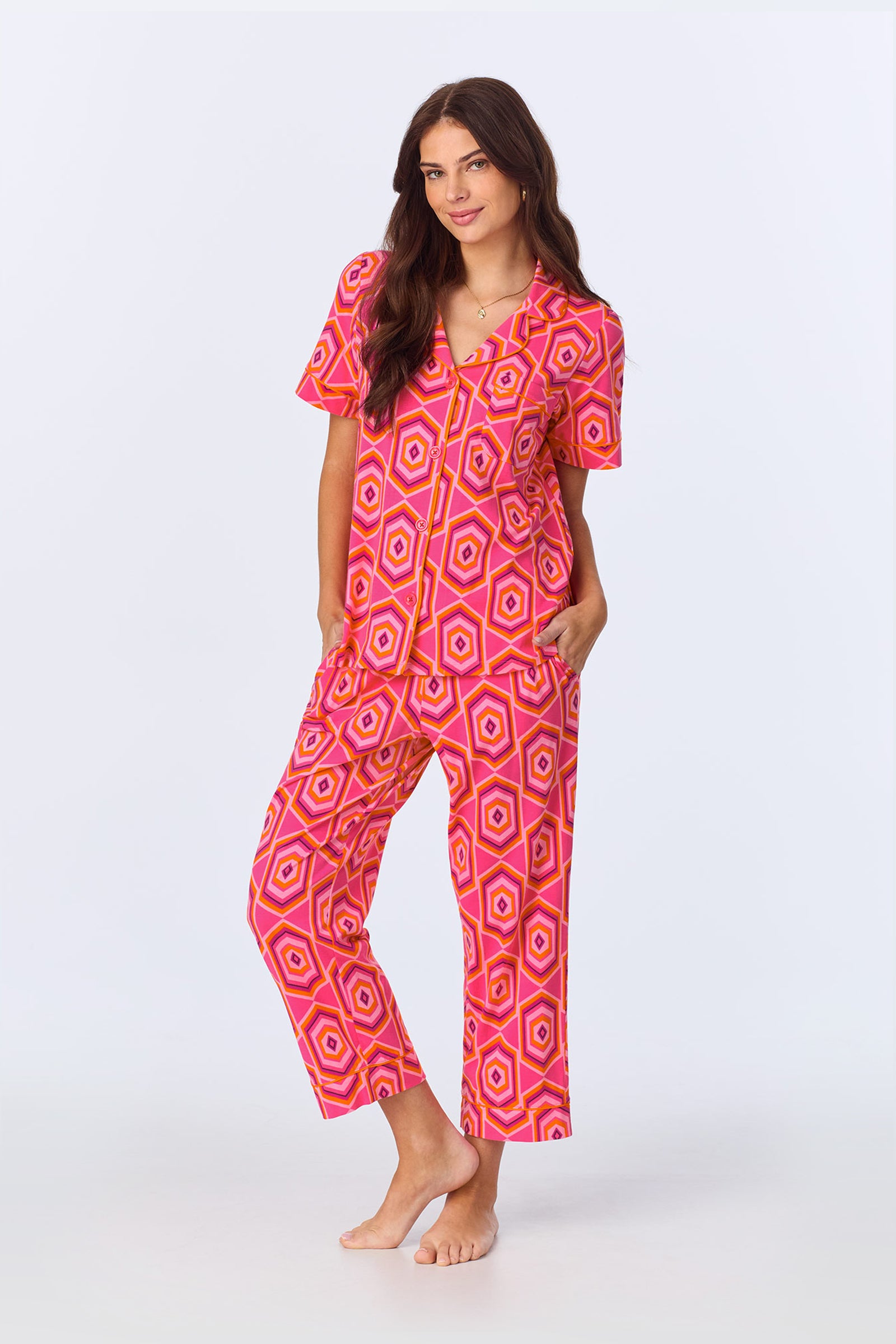 A woman with long brown hair smiles with her hands in her pockets, barefoot and wearing the TT SS Crop PJ Set - MLT Hexagon Jersey featuring a bright pink, orange, and white geometric hexagon pattern against a plain light background.