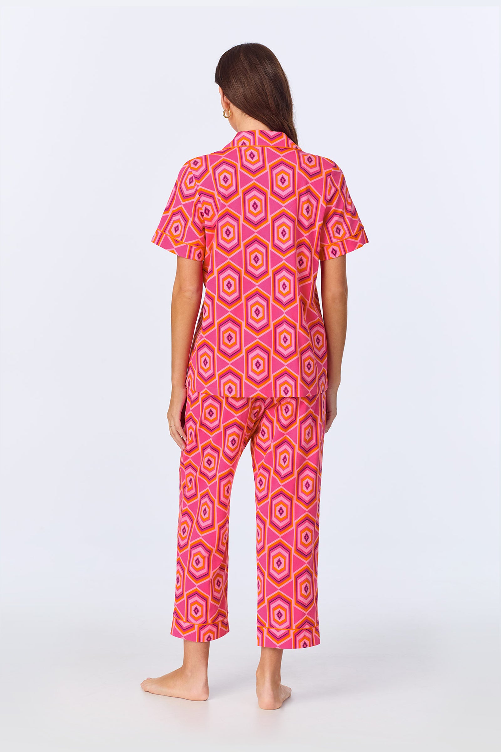 A woman stands barefoot facing away, wearing the TT SS Crop PJ Set - MLT Hexagon Jersey, featuring a matching short-sleeve top and pants with a vibrant multicolor hexagon pattern. The background is plain and light-colored.