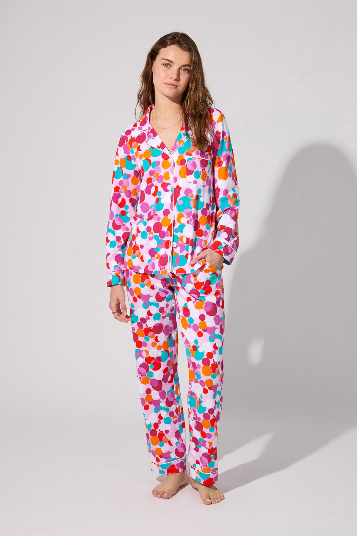 A woman stands barefoot in the Bubble Dots Classic Pj Set—long-sleeved, colorful polka dot pajamas with pants—in shades of pink, red, orange, cyan, and purple against a plain light gray background.