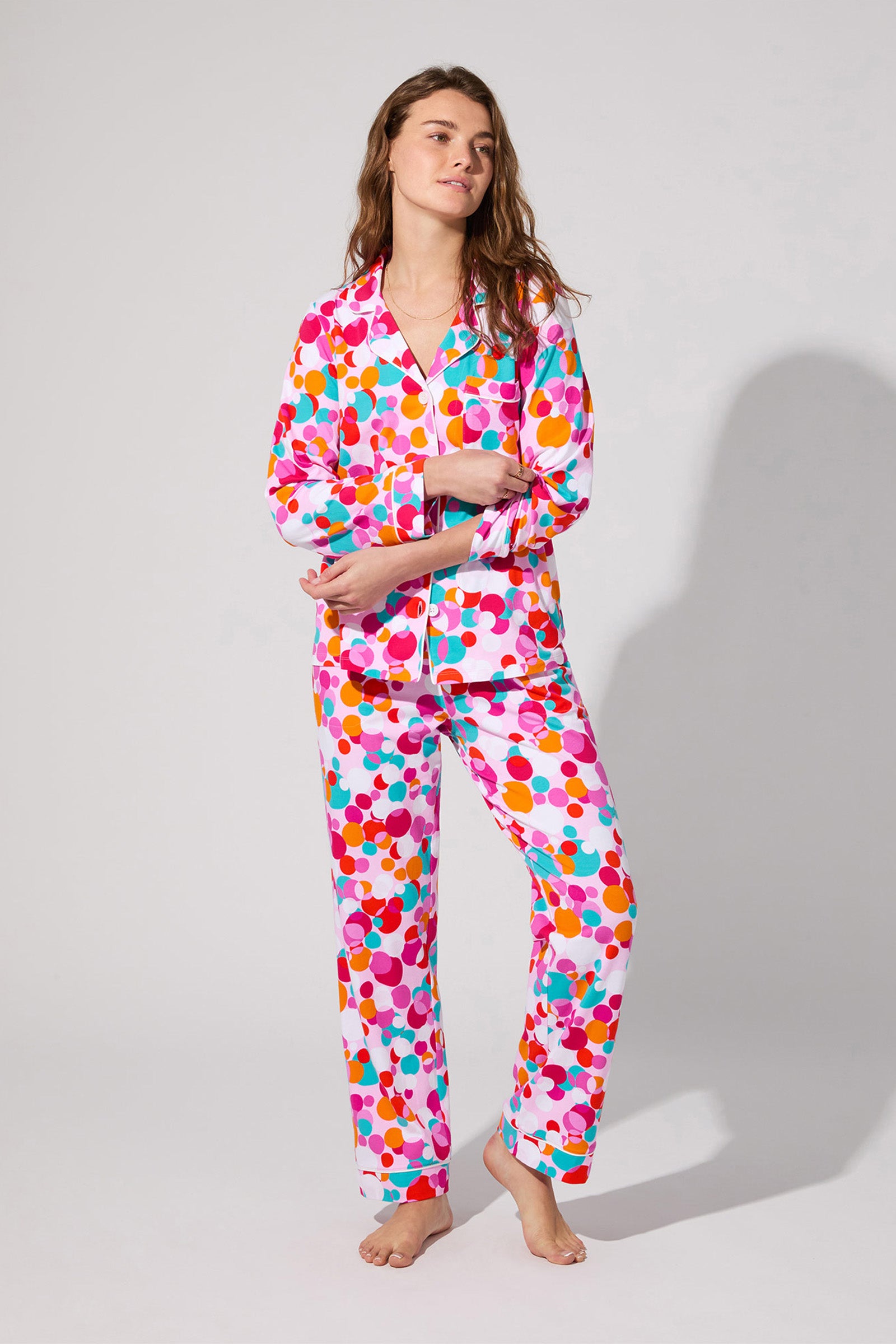 A woman stands barefoot in the Bubble Dots Classic Pj Set, a vibrant pajama with long sleeves and pants featuring pink, orange, red, blue, and white polka dots. She appears relaxed against a plain light background.