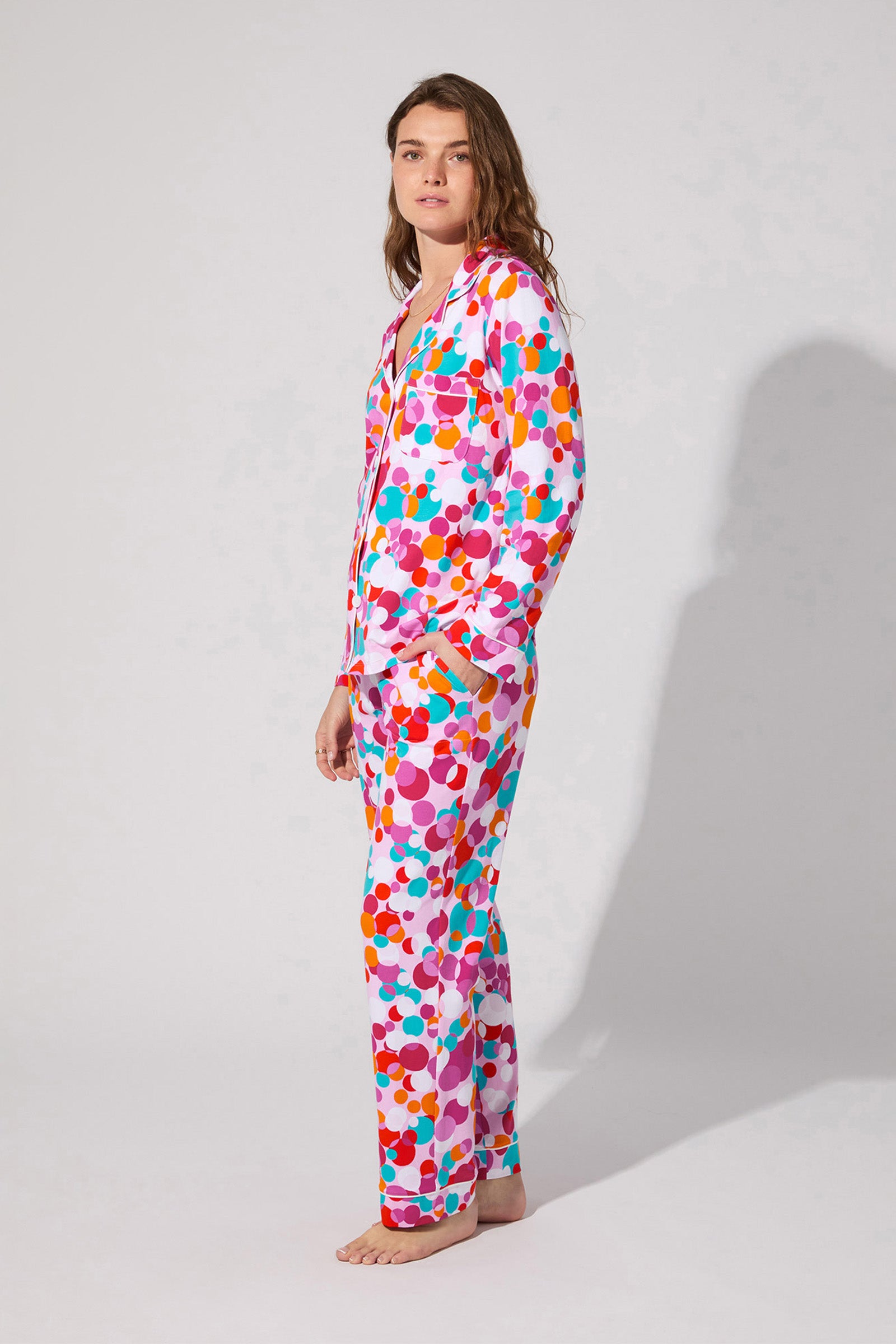 A person stands barefoot, slightly sideways, wearing the Bubble Dots Classic Pj Set featuring a bright, multicolored dot pattern against a plain light background.
