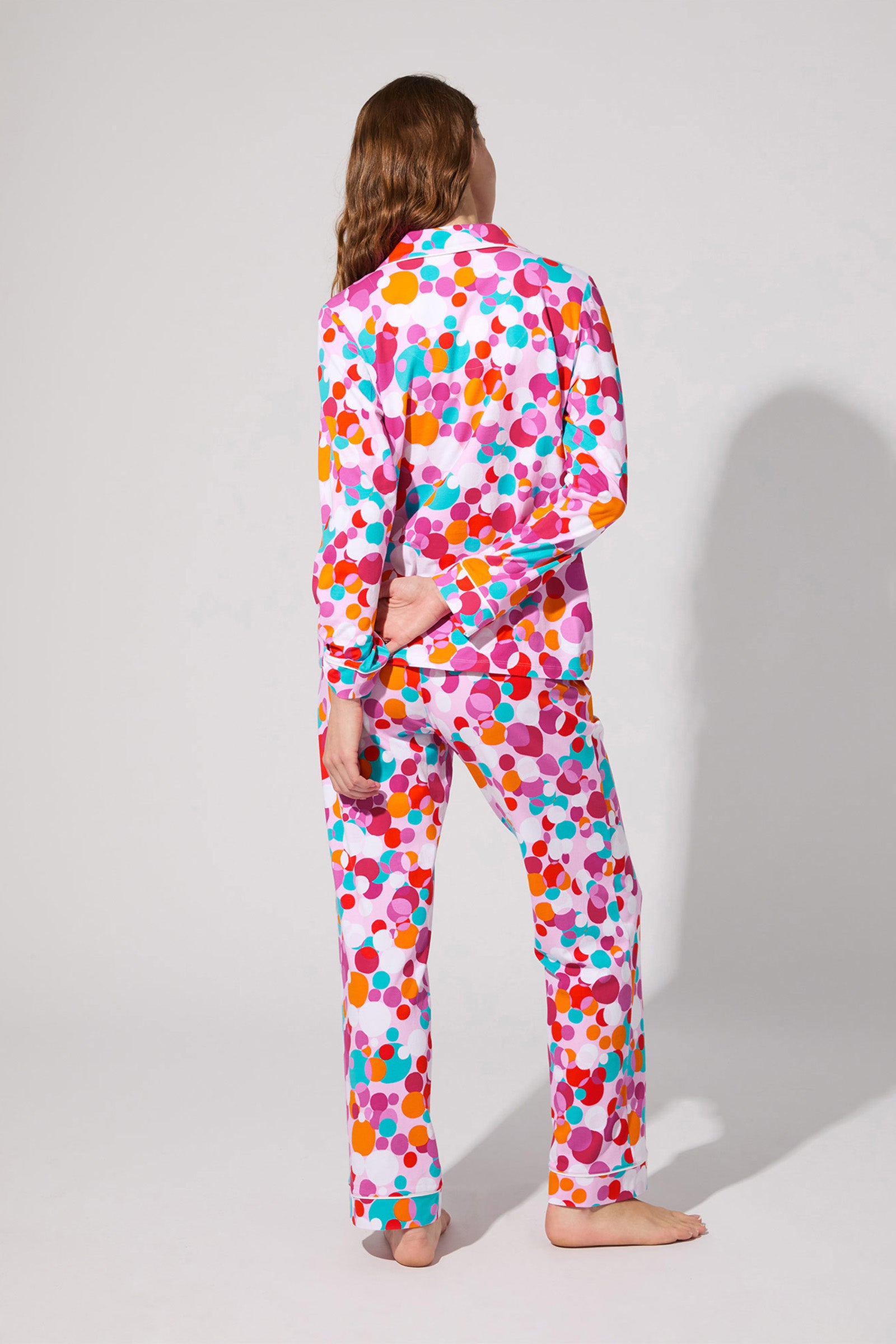 A person with long hair stands barefoot facing away, modeling the Bubble Dots Classic Pj Set—colorful polka-dotted pajamas featuring pink, orange, purple, and blue dots on white—against a plain light gray background.