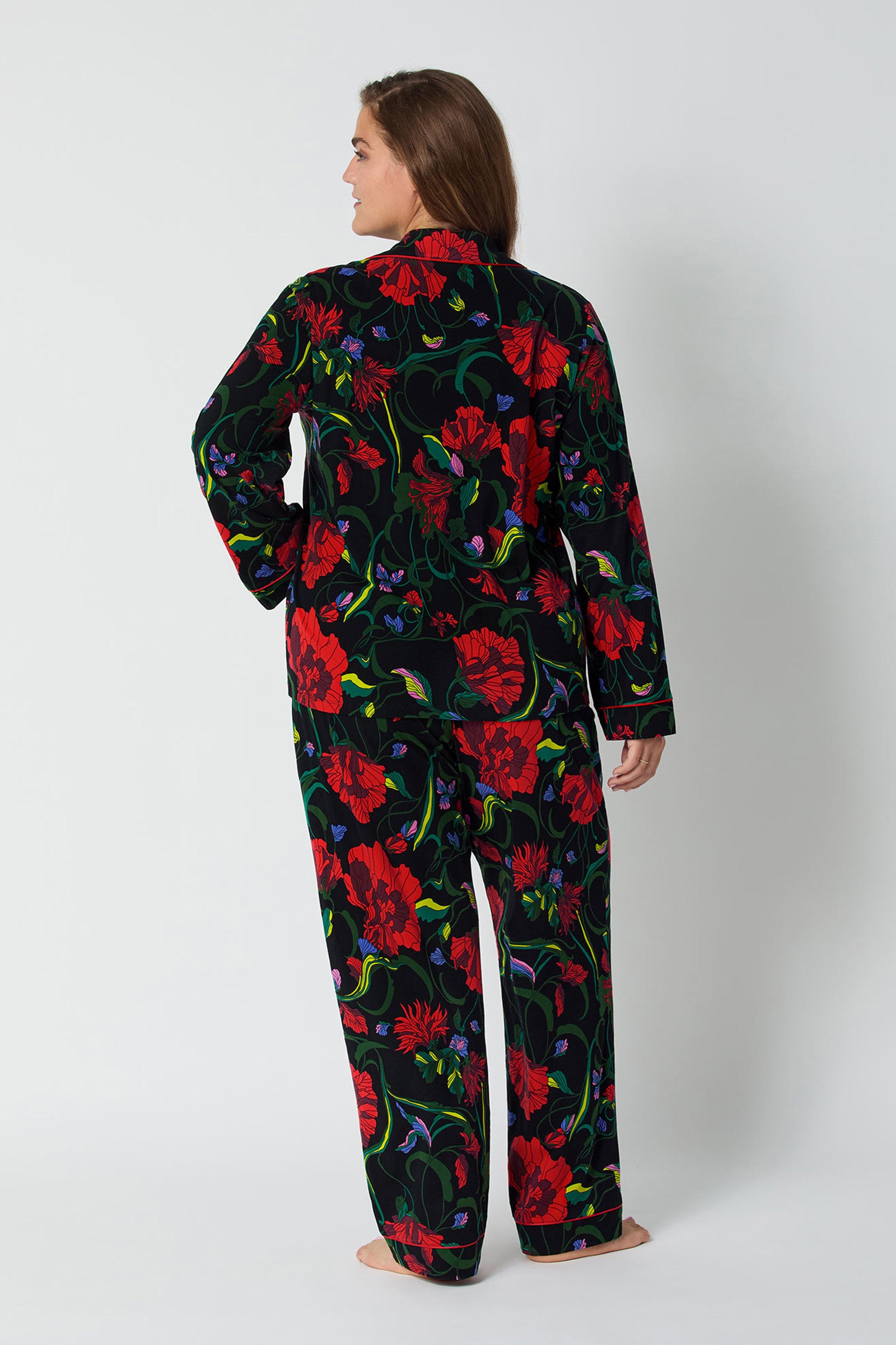 A woman stands barefoot facing away in the Haute Floral L/S Classic Pj—a matching black pajama set with a bold red and green floral pattern—against a plain light background.