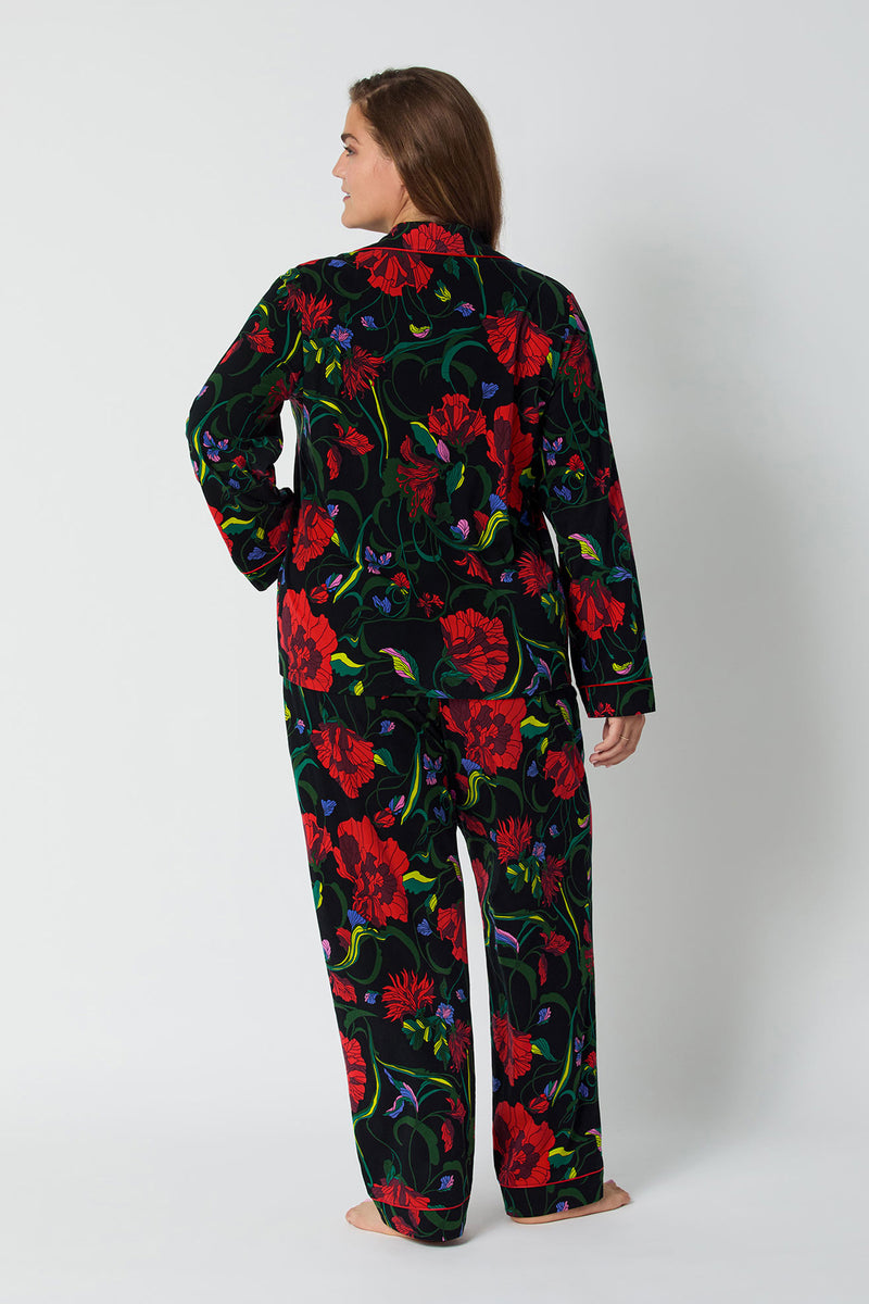 A woman stands barefoot facing away in the Haute Floral L/S Classic Pj—a matching black pajama set with a bold red and green floral pattern—against a plain light background.