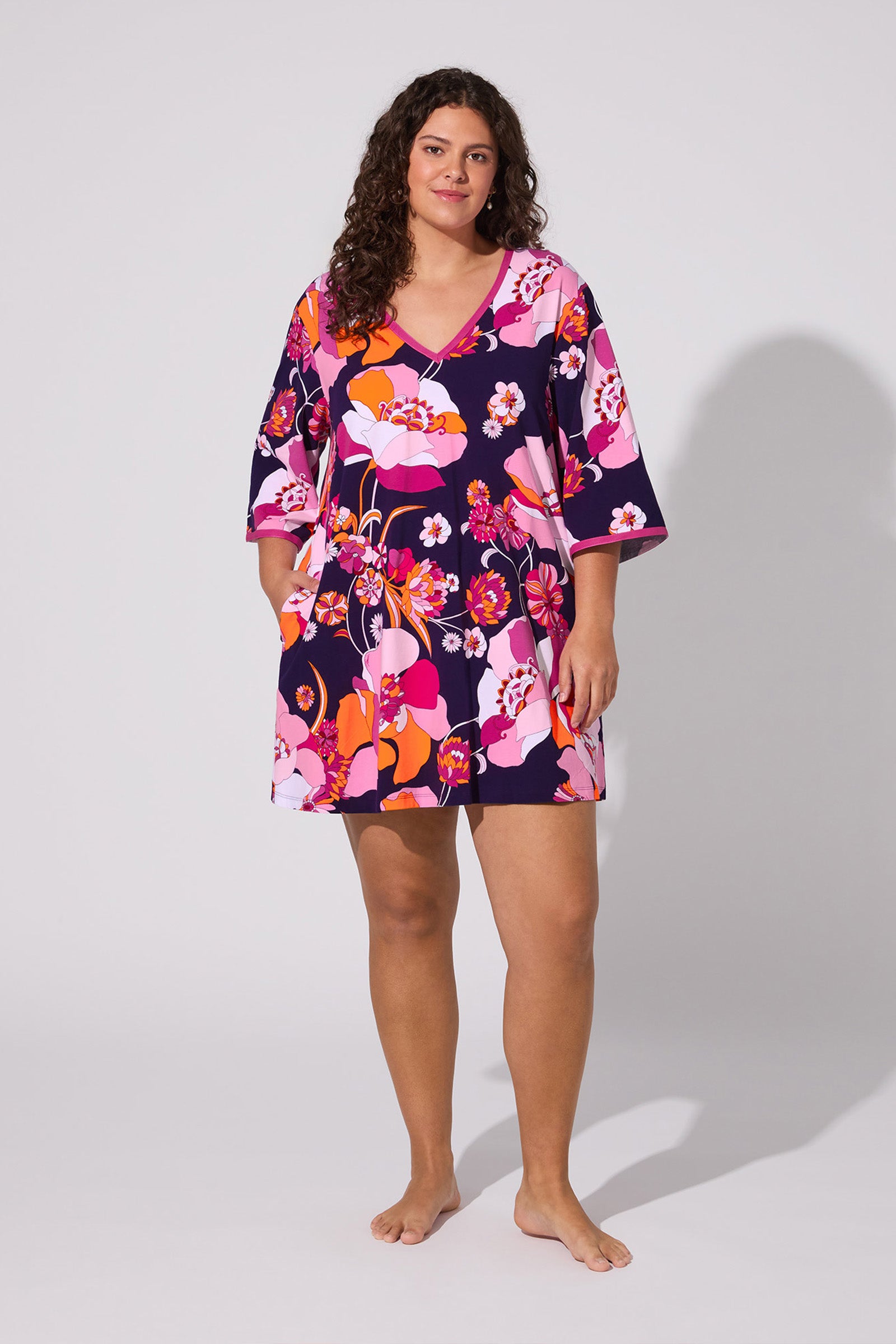 A woman with long curly hair stands barefoot against a plain background, relaxed and confident, wearing the Floral Medley Caftan—a colorful dress with a V-neck and wide sleeves featuring a vibrant floral pattern.
