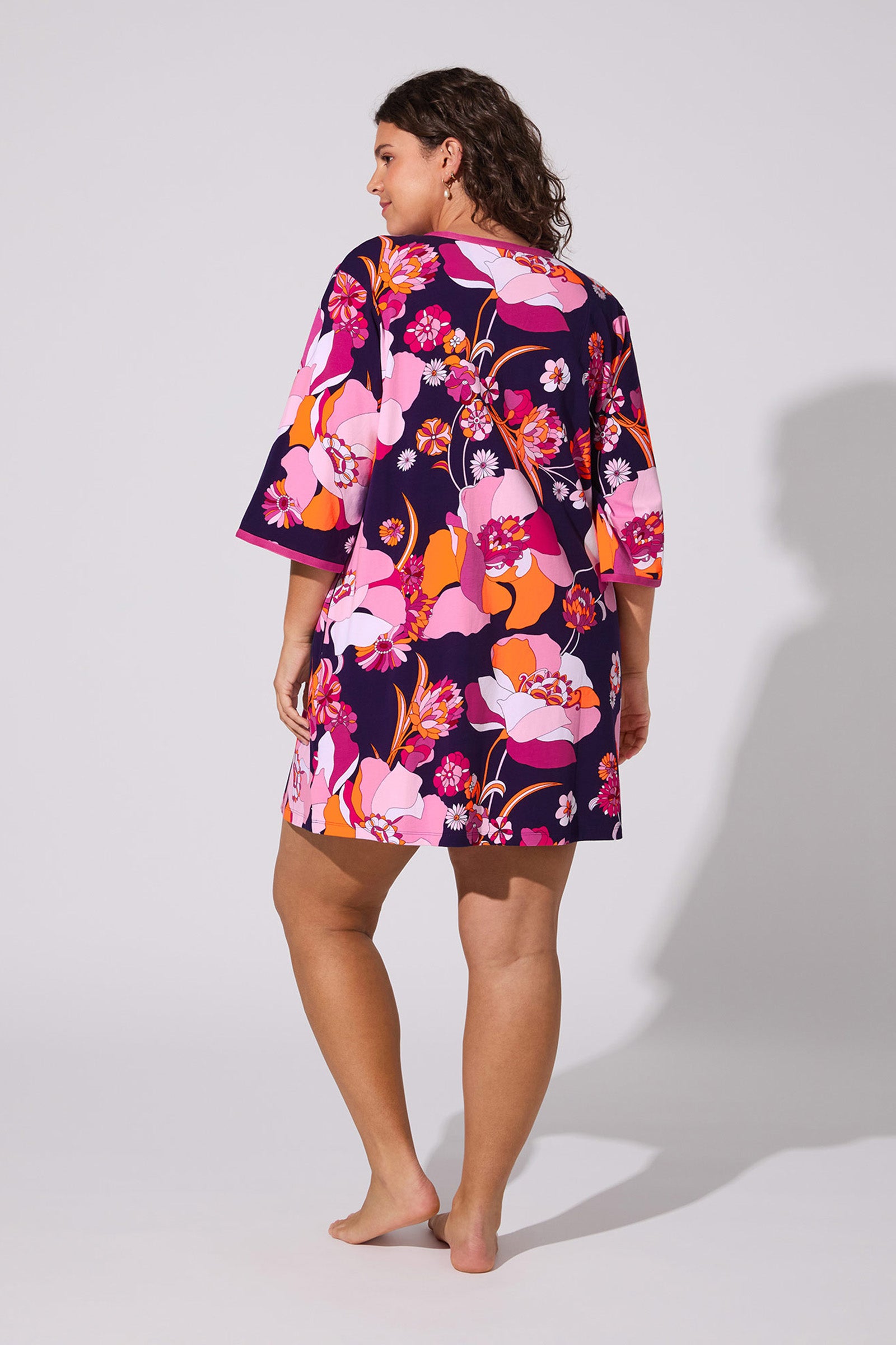 A woman with curly hair stands barefoot, facing away, wearing the Floral Medley Caftan—a vibrant dress with pink, orange, and purple floral patterns—set against a plain light background.