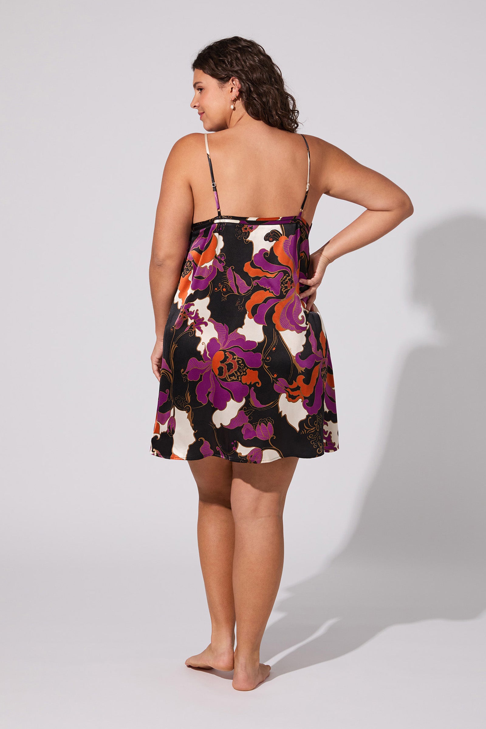 A woman stands barefoot facing away, wearing the Fall Floral Silk Chemise—a sleeveless, knee-length dress with thin straps and a bold floral pattern in purple, orange, black, and white. The background is plain and light-colored.