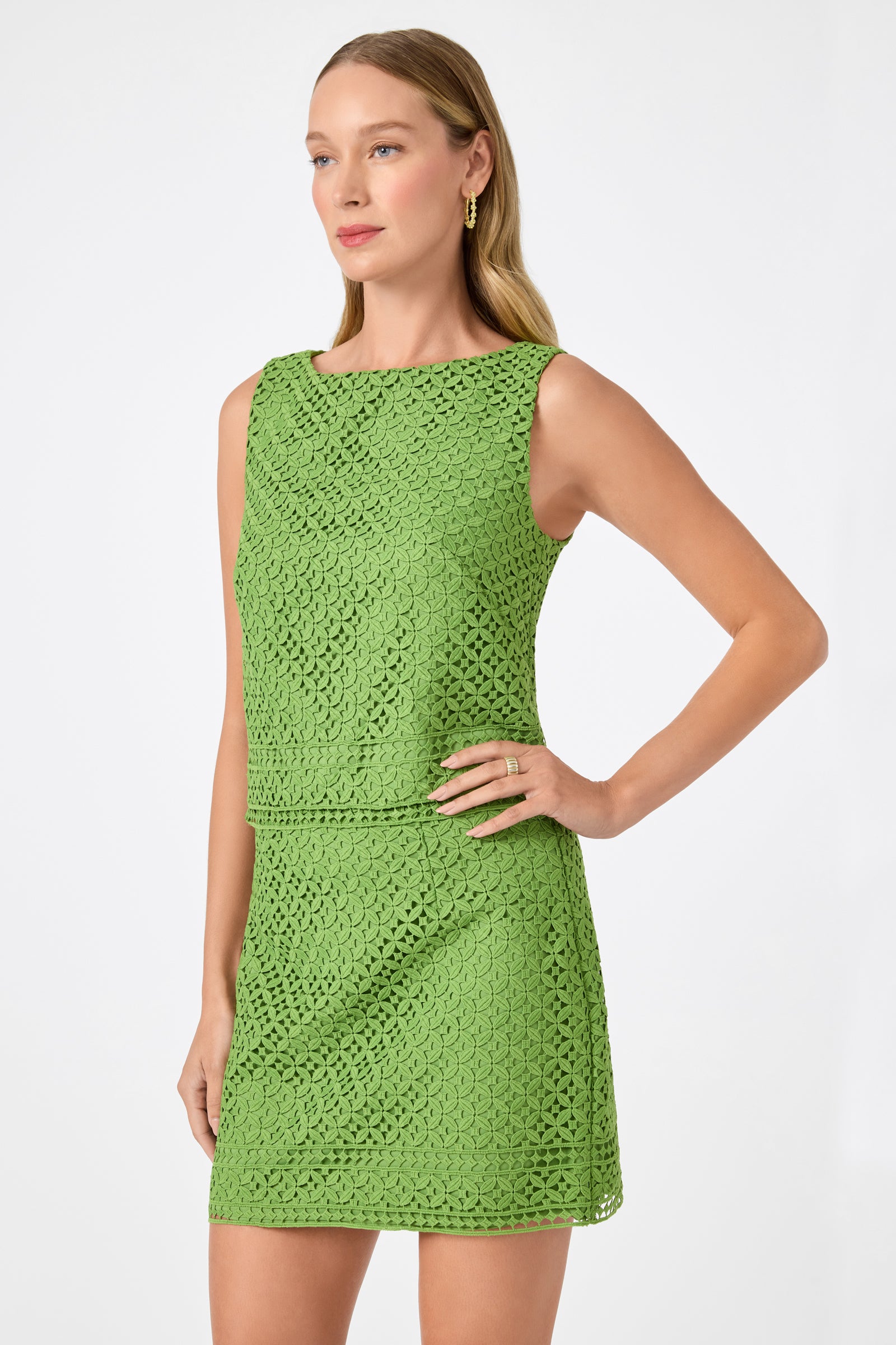 A woman with long blonde hair and gold hoop earrings wears the Cayley 2 Top in Lumi Lace, a sleeveless green crochet-patterned piece, standing with one hand on her hip against a plain white background.