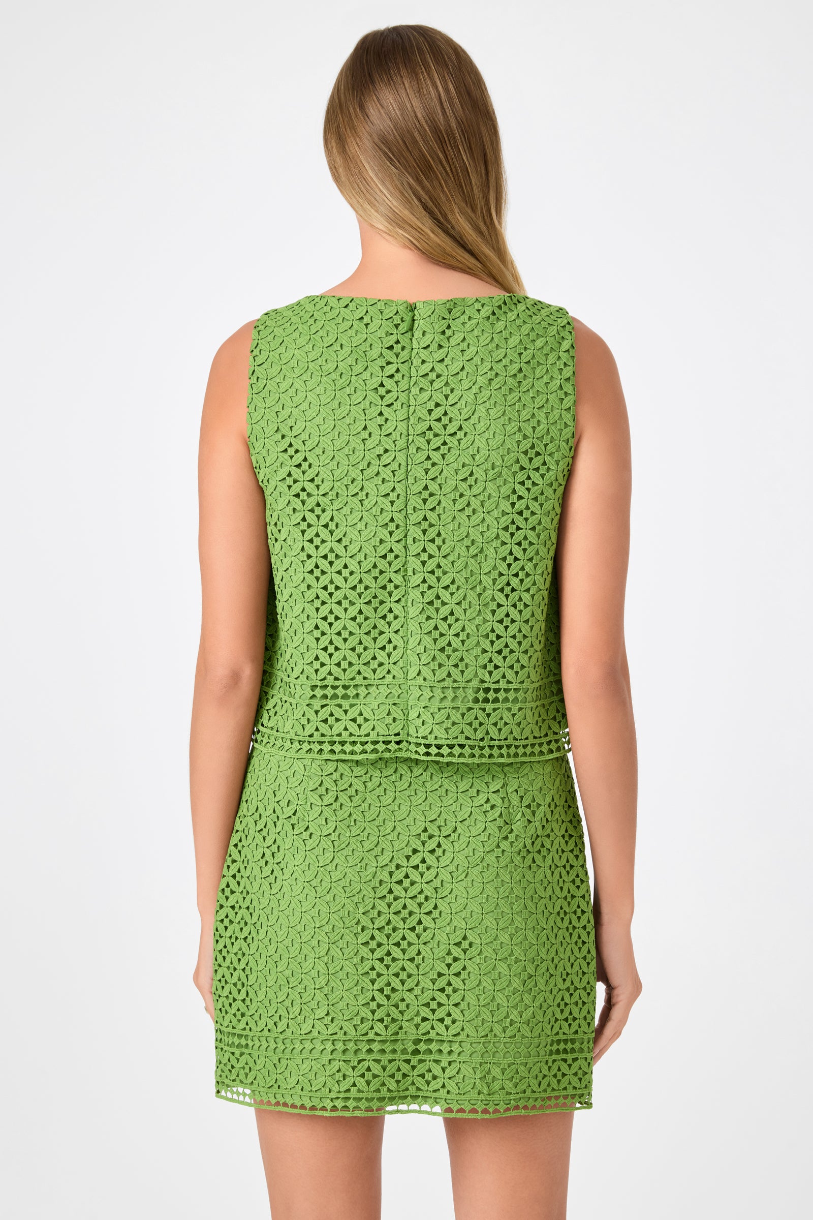 A woman faces away, wearing the Cayley 2 Top - Lumi Lace, a sleeveless green lace top with a fitted waist and short hem. Her straight light brown hair falls past her shoulders against a plain white background.