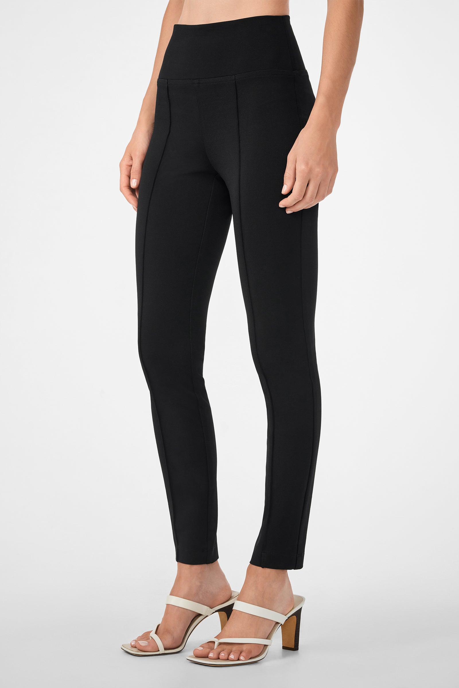 A person wears the Rose Pant high-waisted black leggings with white heeled sandals, standing against a plain light background. The image is cropped from the waist down to highlight the clothing and footwear.