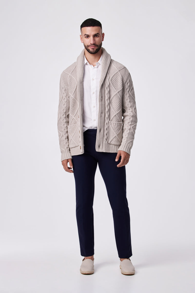A man faces forward against a plain white background, wearing a beige cable-knit cardigan, white button-up shirt, and Clyde Slim Trouser - Stretch Piqué in dark navy with beige slip-on shoes.