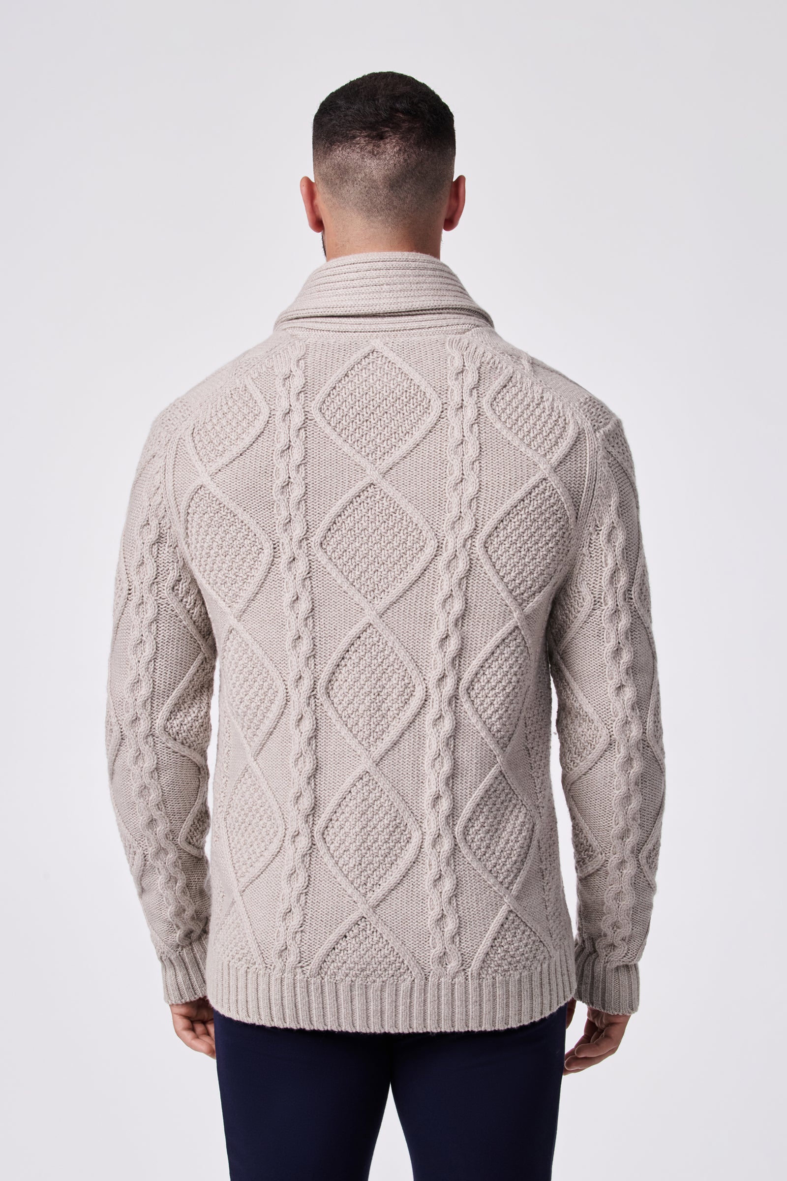 A person with short dark hair is seen from behind wearing the Brighton Cardigan—a light beige, cable-knit sweater featuring diamond and braid patterns, plus ribbed hem and collar—against a plain white background.