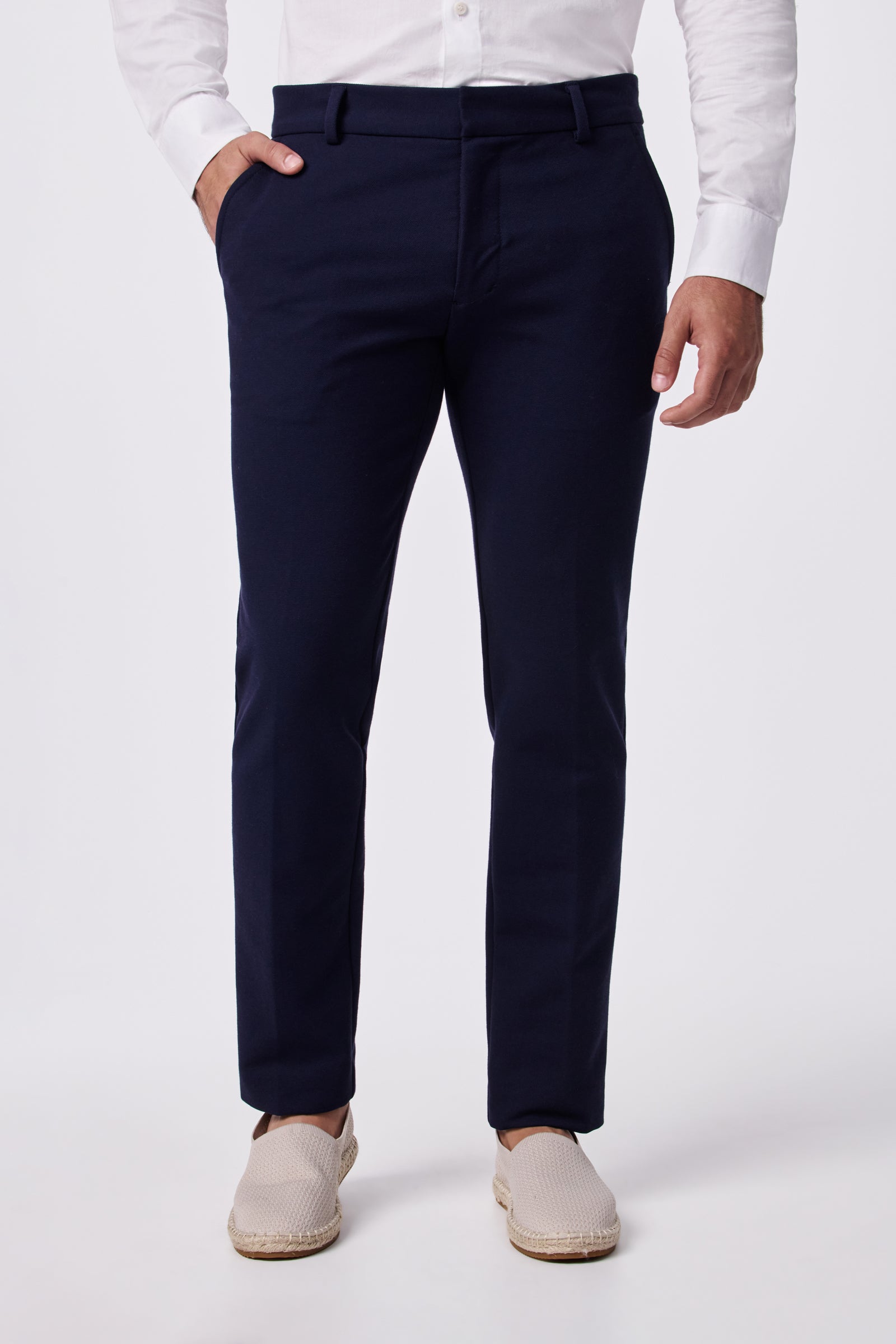 A man wearing the Clyde Slim Trouser - Stretch Piqué in navy blue, paired with a white dress shirt and beige slip-on shoes, stands with one hand in his pocket against a plain white background.