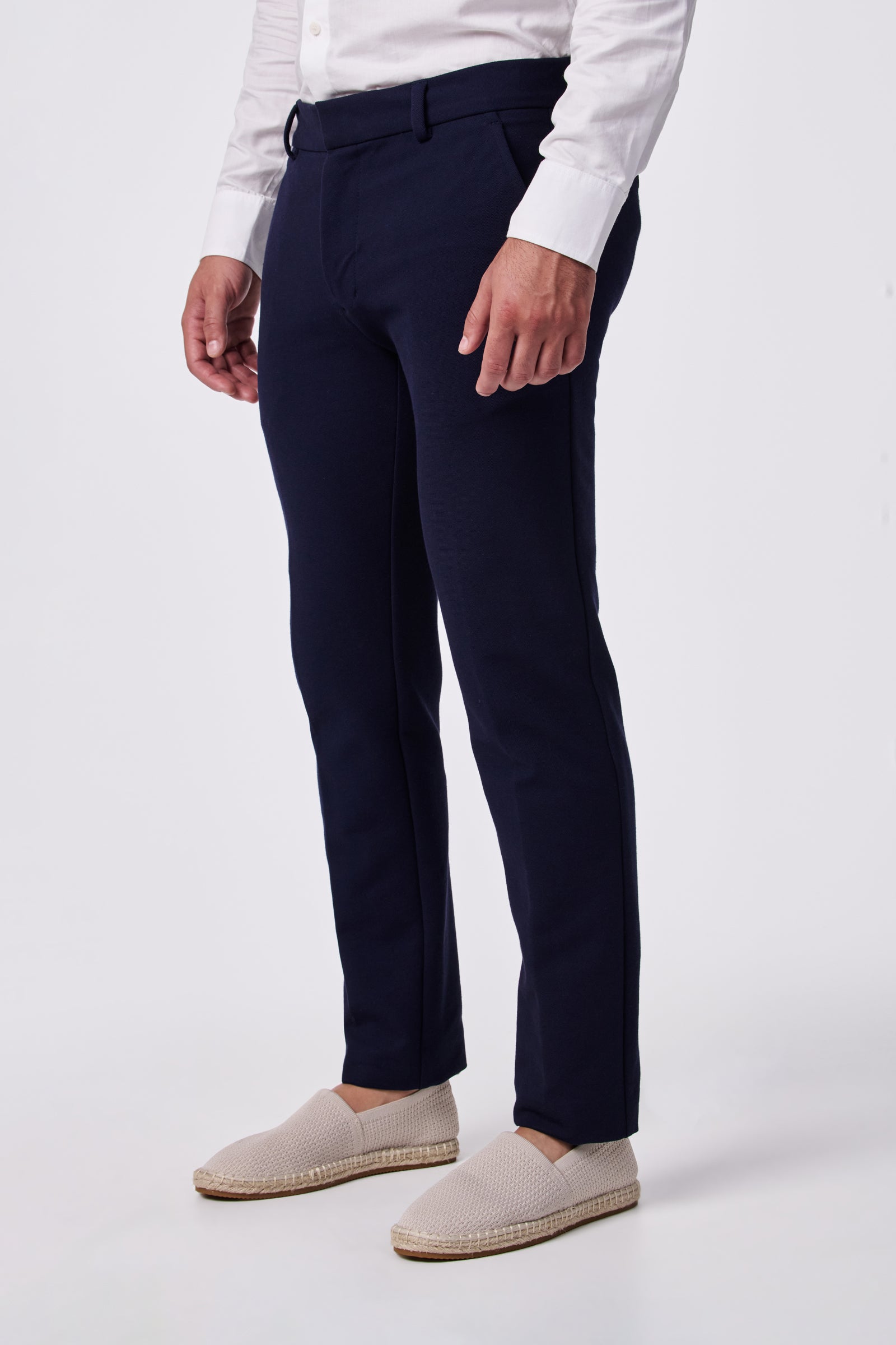 Wearing the Clyde Slim Trouser - Stretch Piqué in dark blue, a person stands against a white background, highlighting the lower body and legs. They pair the trousers with a white long-sleeve shirt and light slip-on shoes.