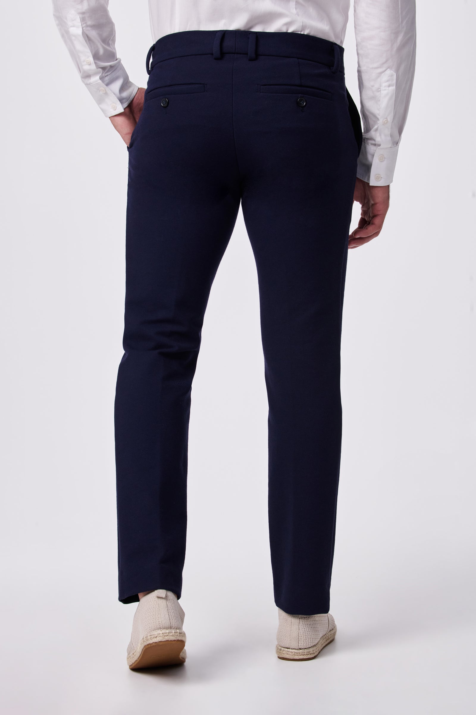 Shown from the back against a plain white background, a person wears navy blue Clyde Slim Trouser - Stretch Piqué paired with a white dress shirt and light-colored textured shoes.