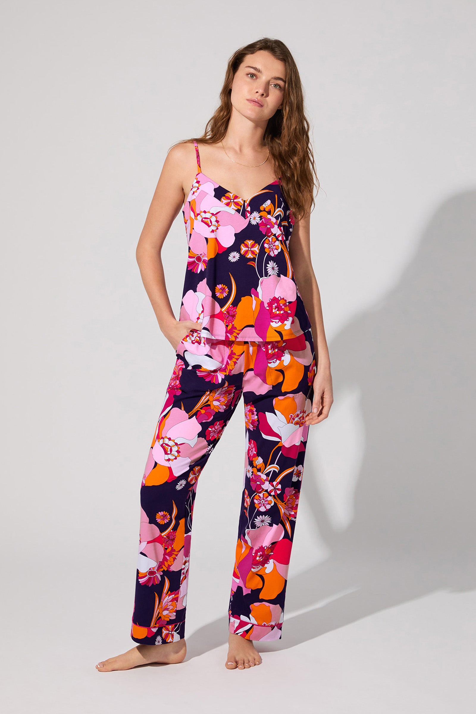 A woman with long, wavy hair stands barefoot in the Floral Medley Cami Pj Set, featuring a colorful floral pattern in pink, red, orange, and purple. The set includes a sleeveless V-neck top and loose pants; she looks relaxed.