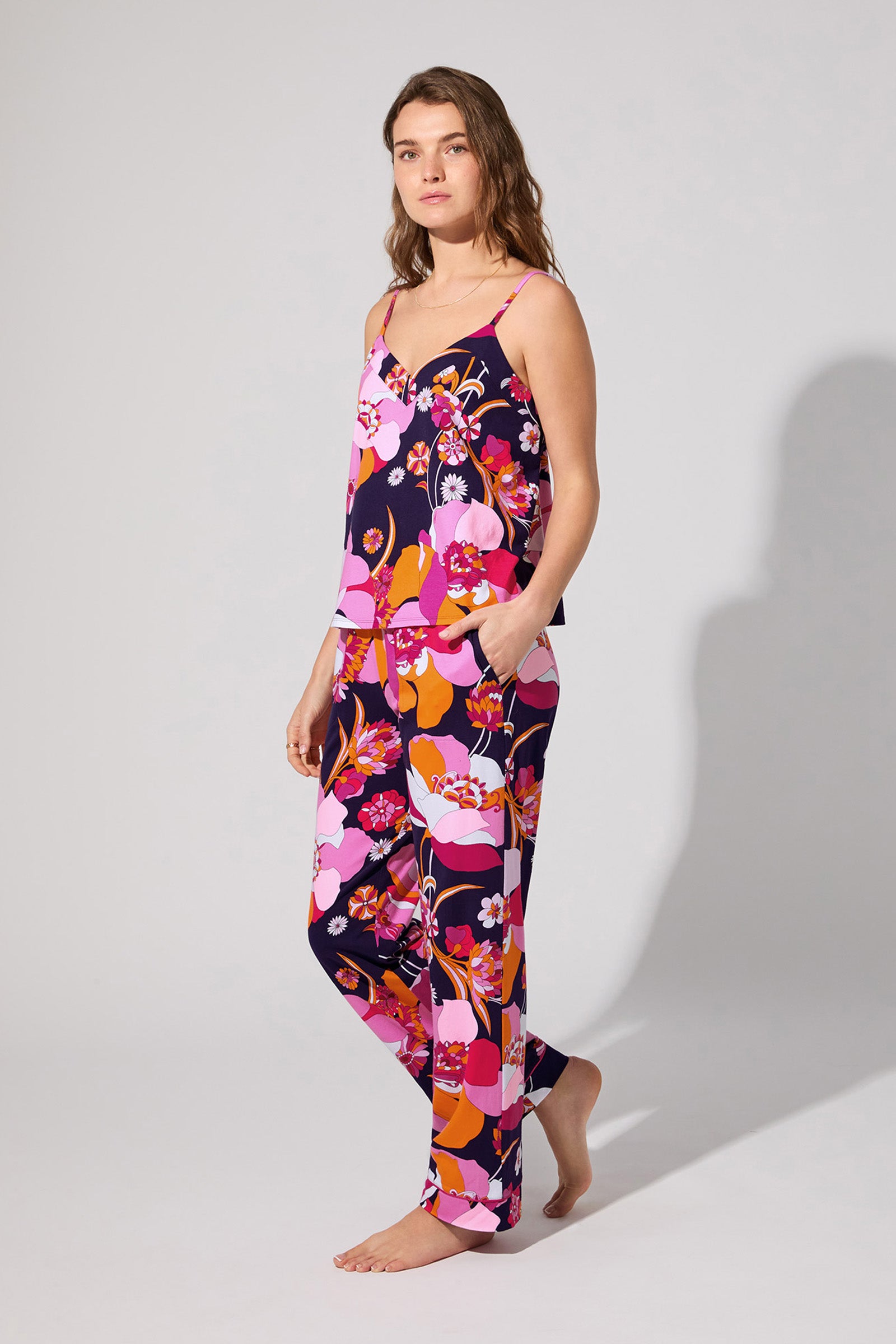 A woman stands barefoot in the Floral Medley Cami Pj Set, featuring a colorful floral print, sleeveless top, and long pants. She poses against a plain background with a relaxed expression, looking slightly to the side.