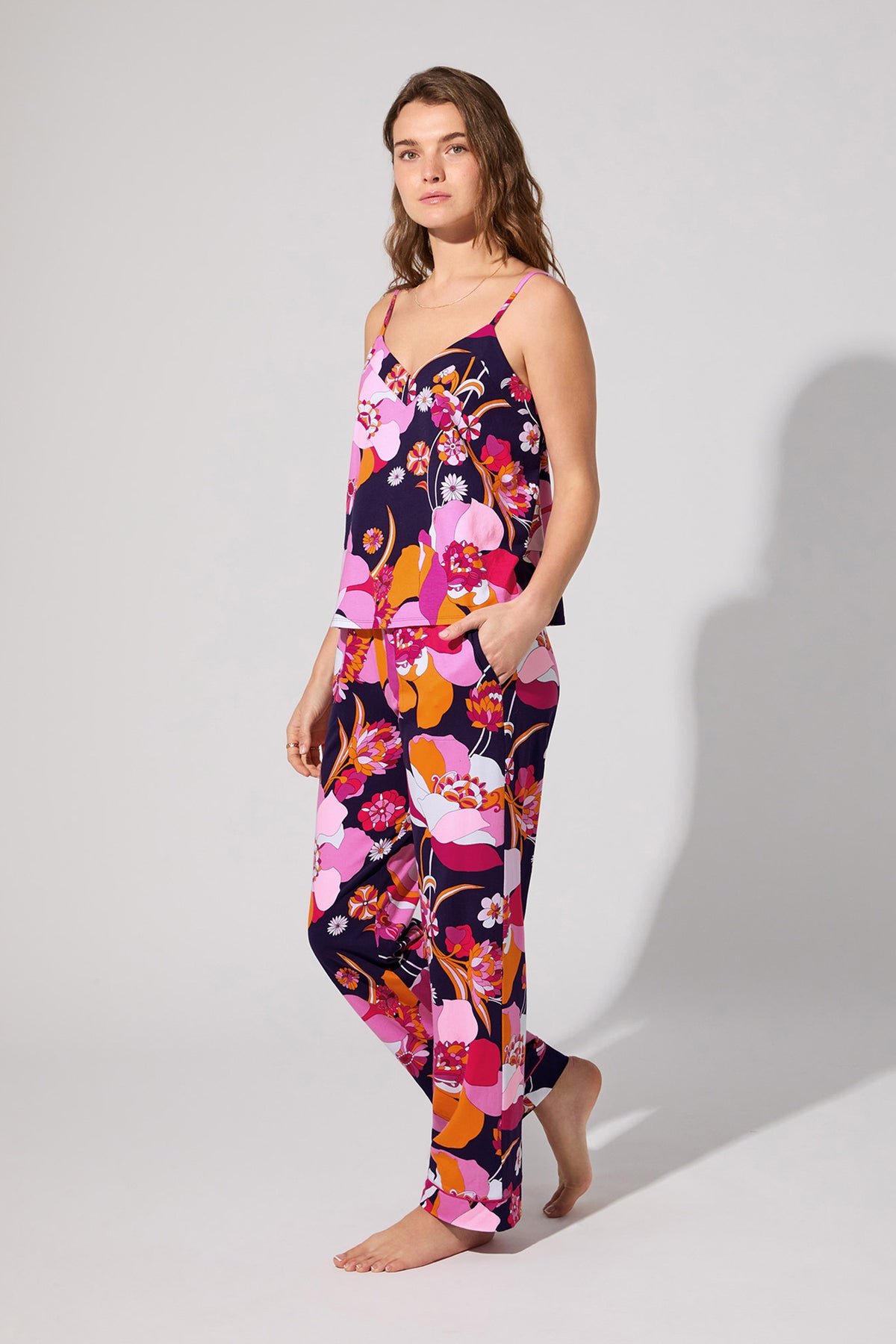 A woman stands barefoot in the Floral Medley Cami Pj Set, featuring a colorful floral print, sleeveless top, and long pants. She poses against a plain background with a relaxed expression, looking slightly to the side.