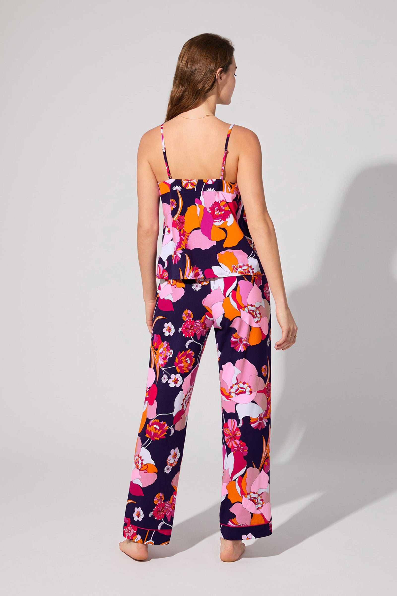 A woman stands barefoot facing away, wearing the Floral Medley Cami Pj Set with a loose camisole and wide-leg pants in vibrant pink, orange, red, and purple florals. The background is plain and light gray.